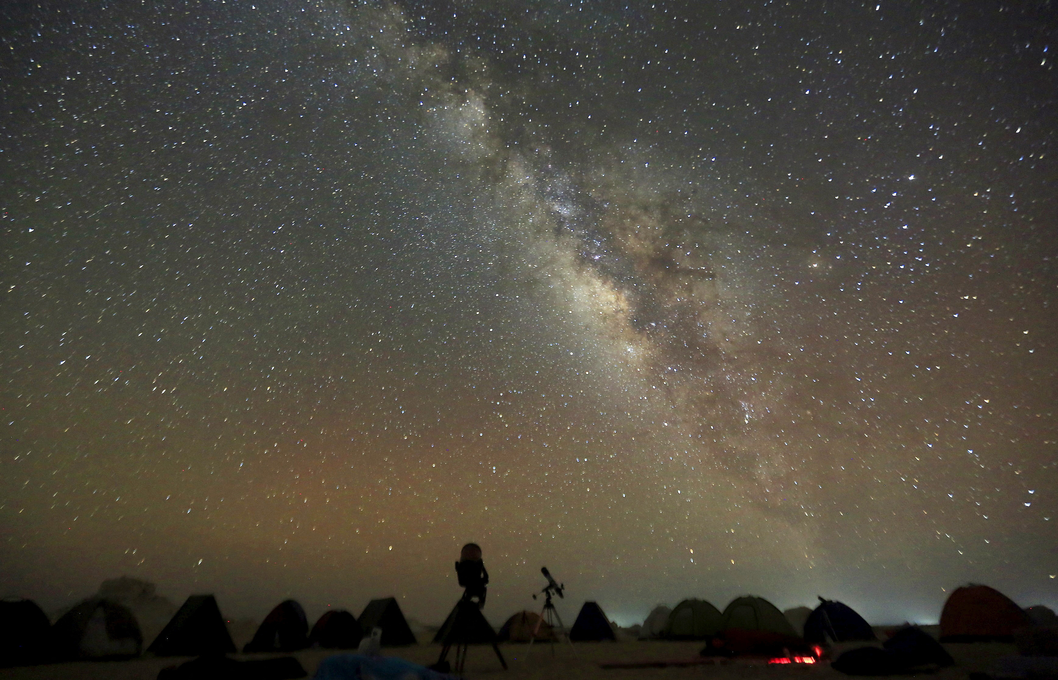 The Milky Way is seen in the night sky around telescopes and camps of people over rocks in the White Desert north of the Farafra Oasis southwest of Cairo