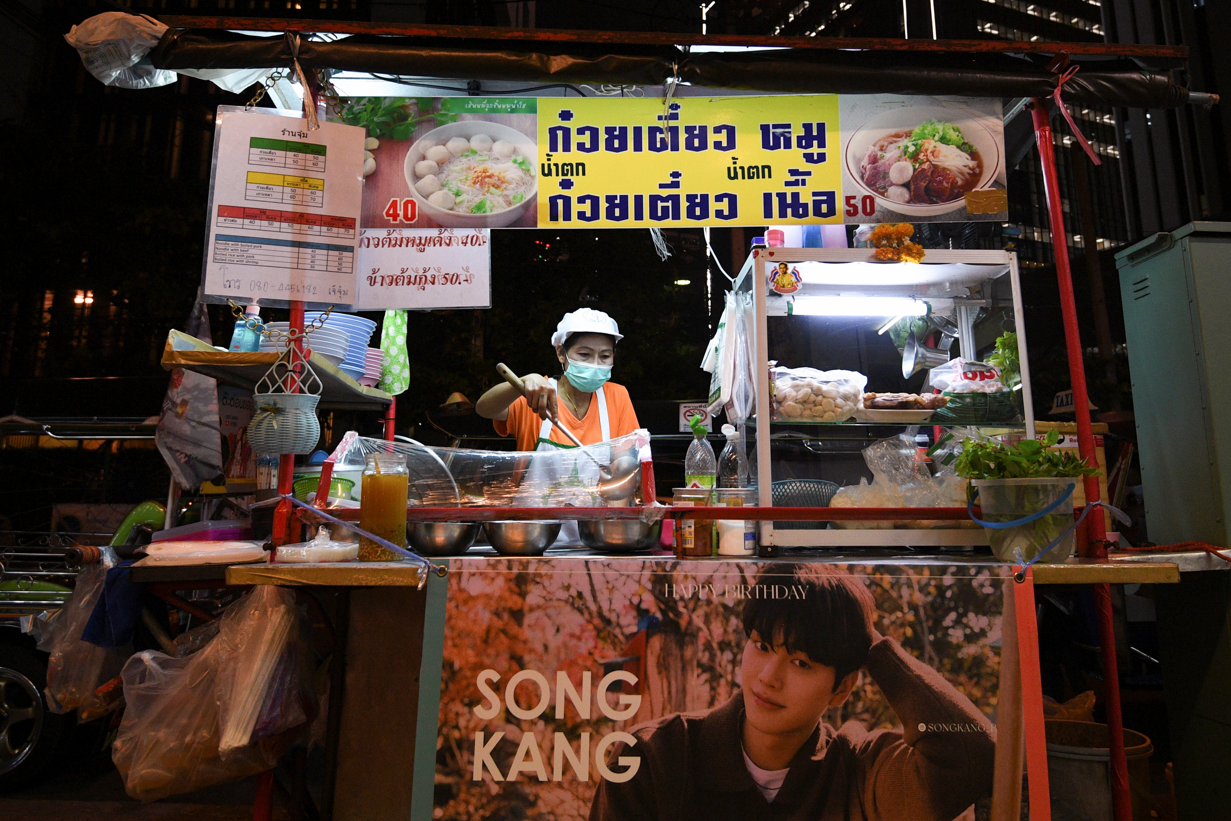 A woman selling street food has her stall decorated with a banner of Korean star Song Kang in Bangkok