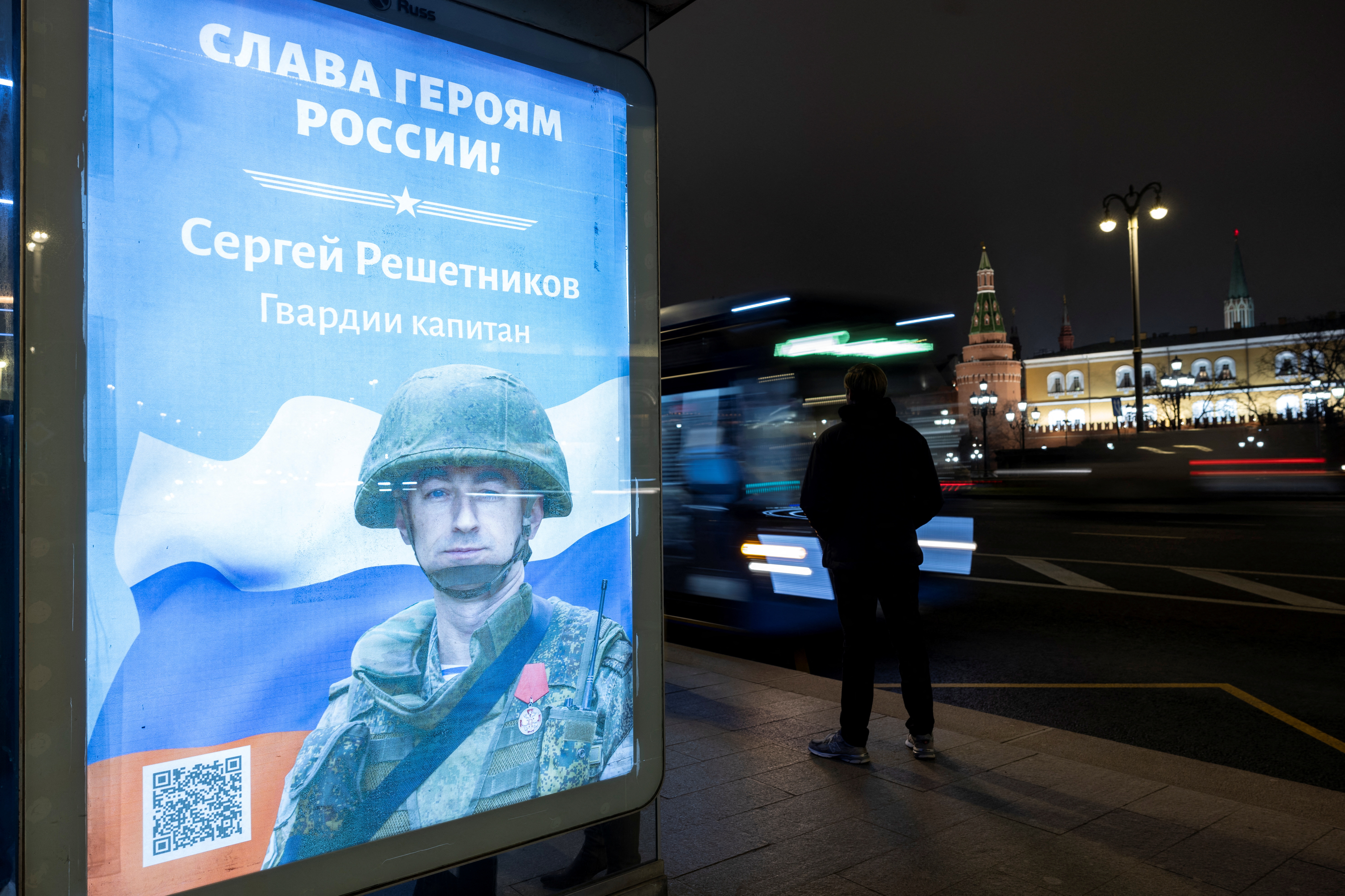 A person waits for a bus at a bus stop with a board displaying an image of Russian service member in Moscow