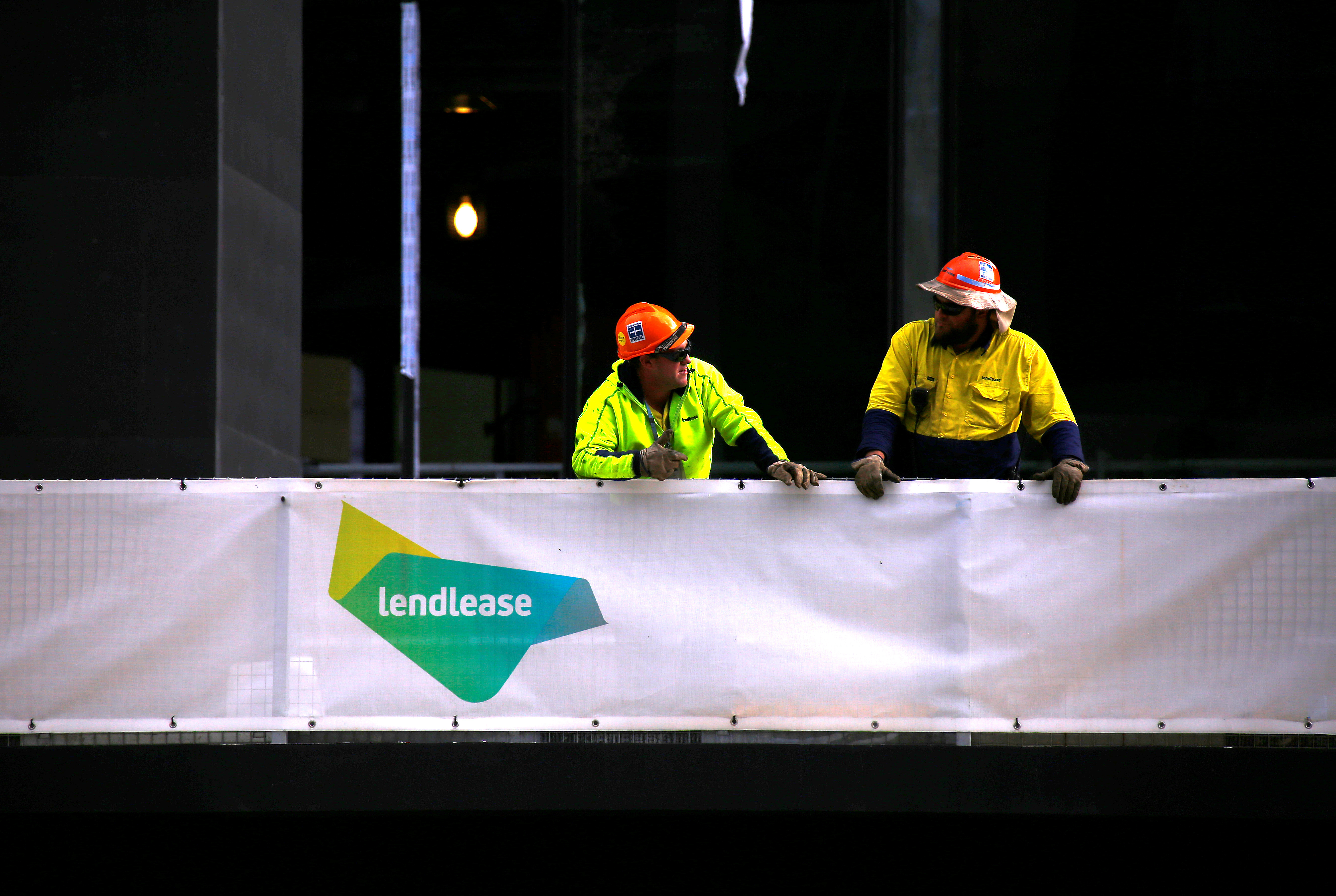 Construction workers lean on a fence adorned with a sign for the construction company Lendlease at a construction site in central Sydney, Australia