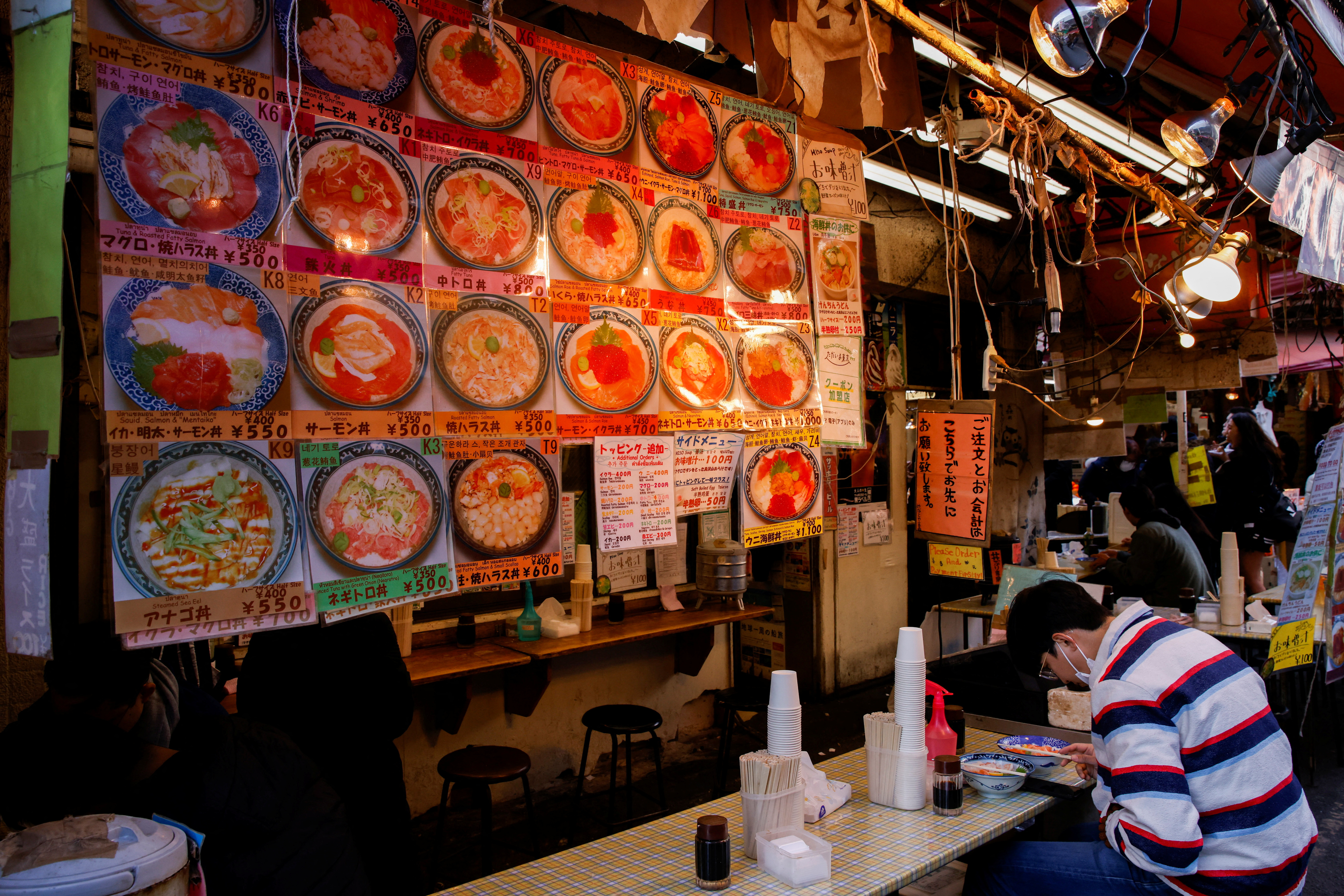 Man eats under menu pictures with prices, at a restaurant of a market in Tokyo