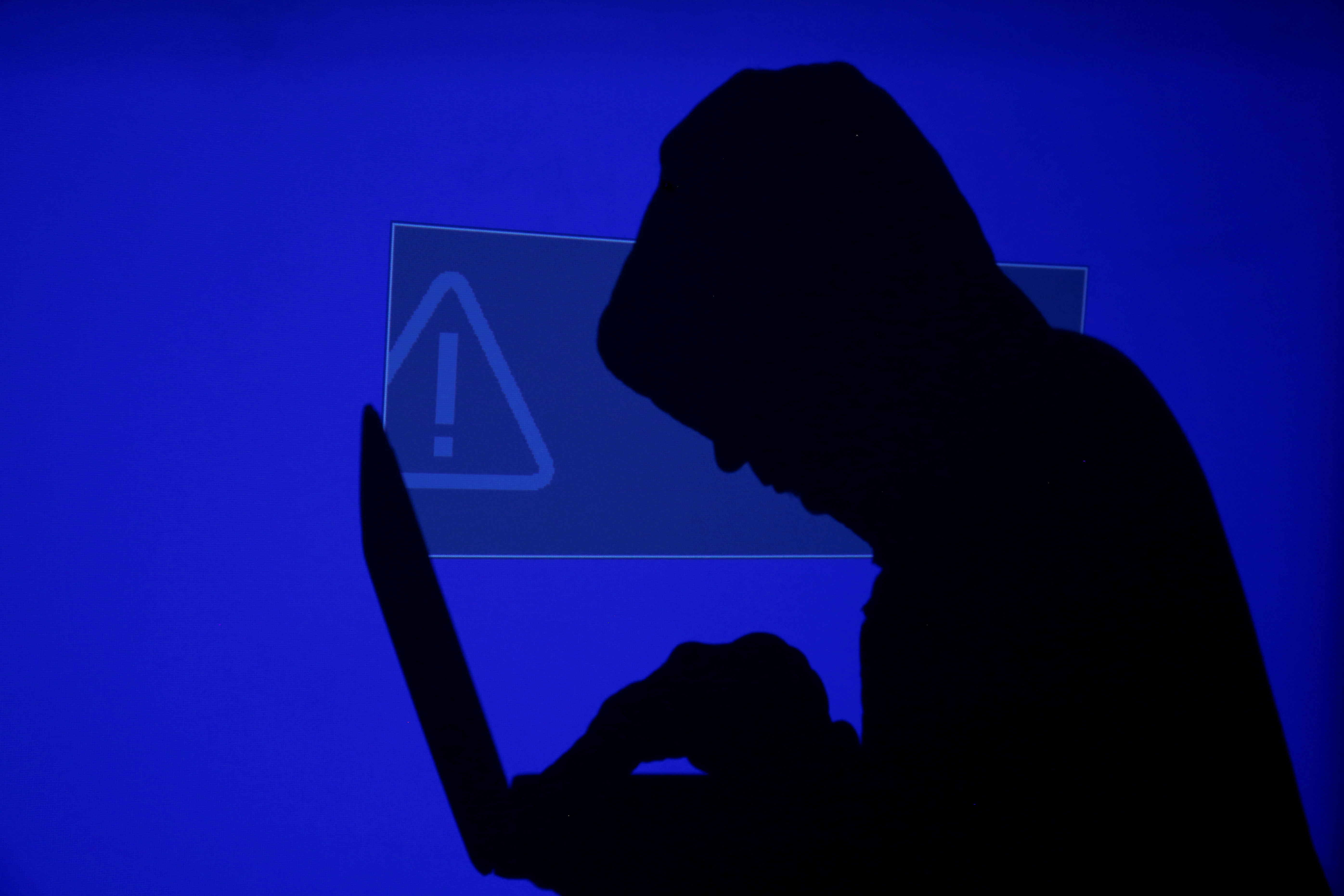 Hooded man holds laptop computer as blue screen with an exclamation mark is projected on him in this illustration picture