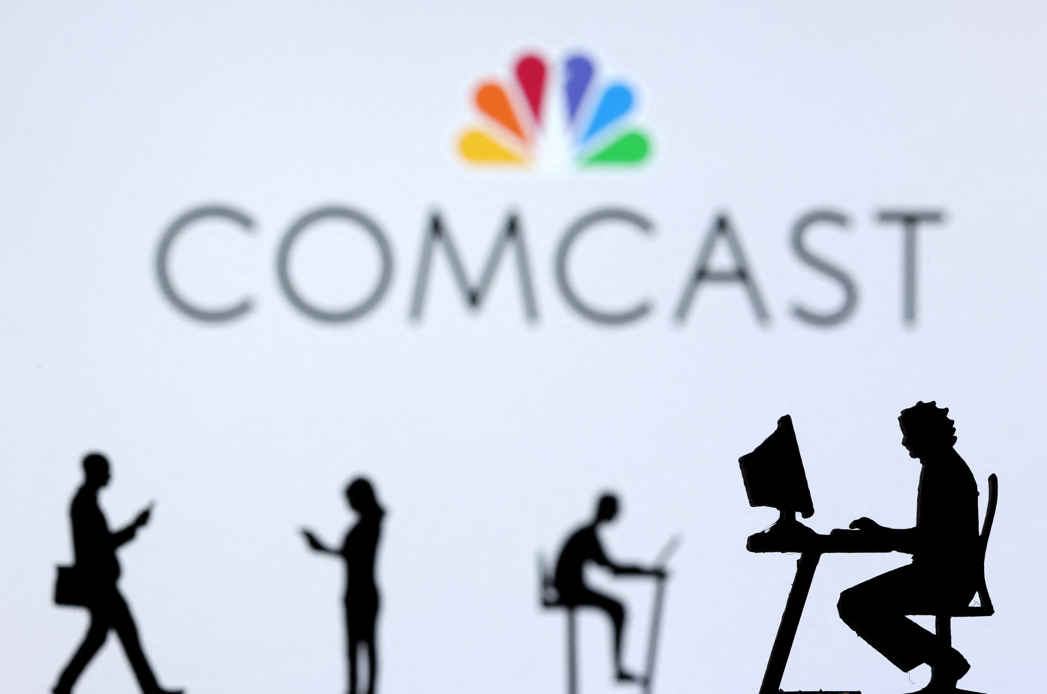 Illustration shows small toy figures with laptops and smartphones in front of displayed Comcast logo