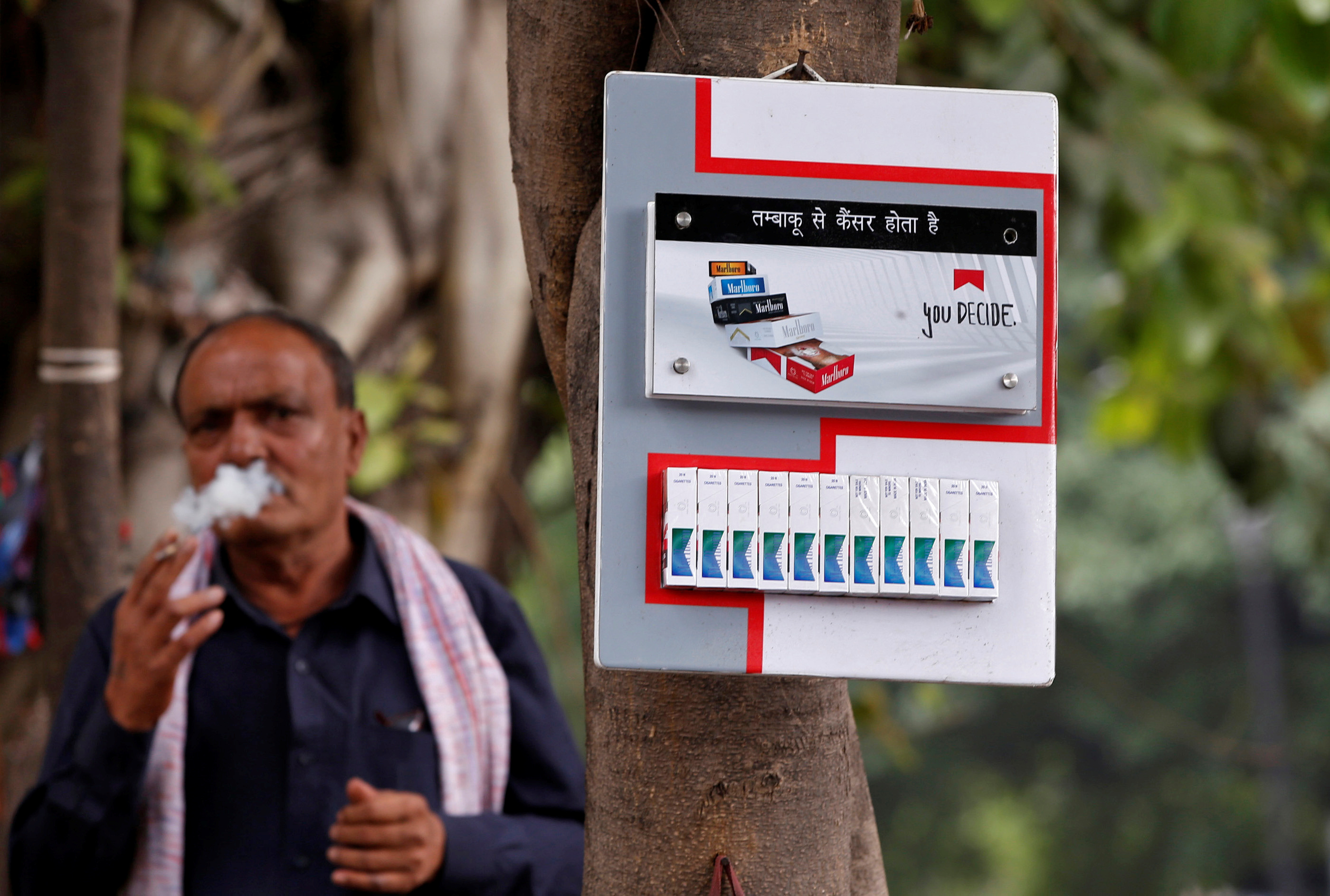 A man smokes next to a cigarette advertisement hung on a tree at a marketplace in New Delhi