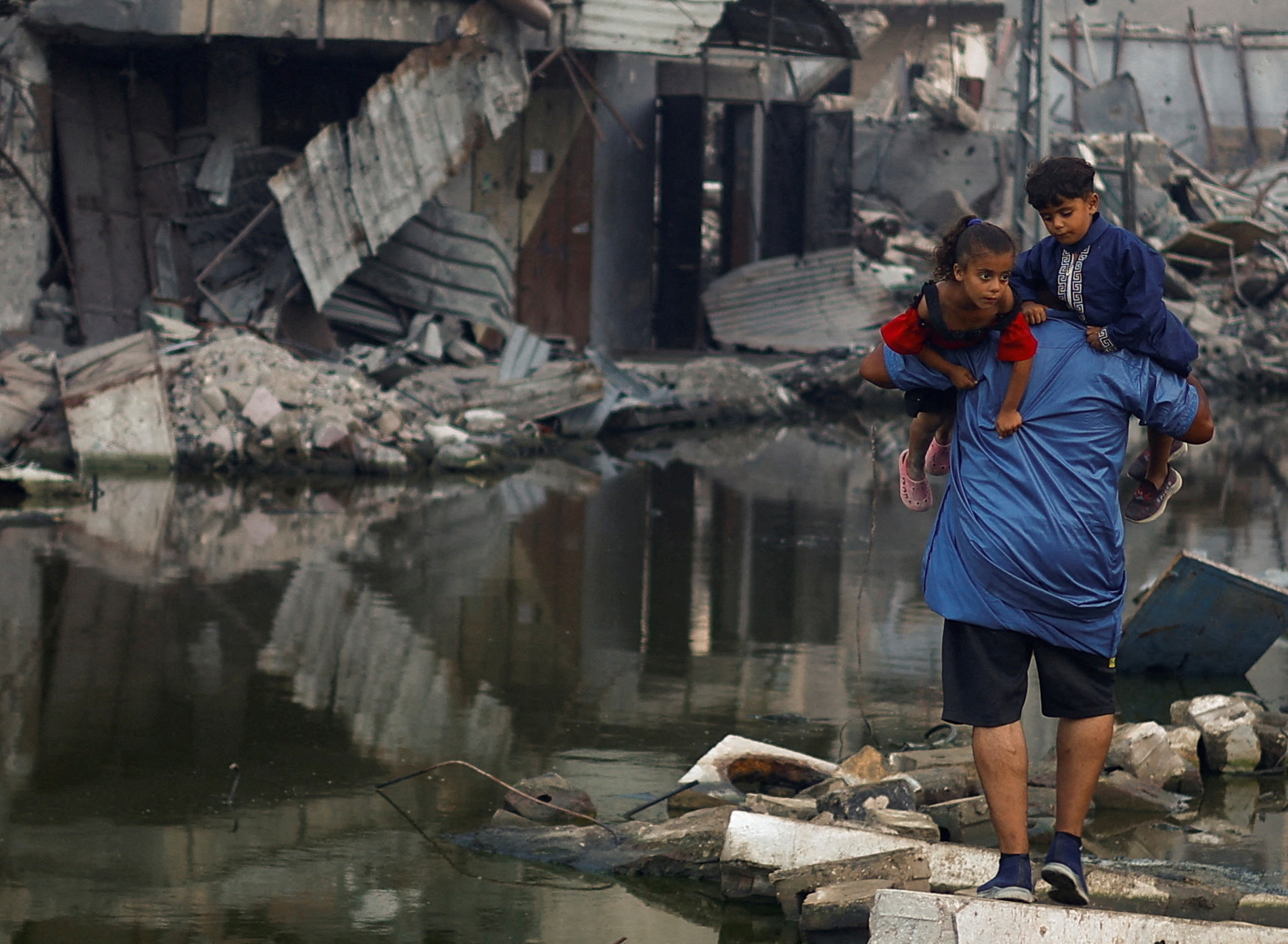 A Palestinian man holds his children as he walks next to buildings destroyed in an Israeli strike, in Khan Younis