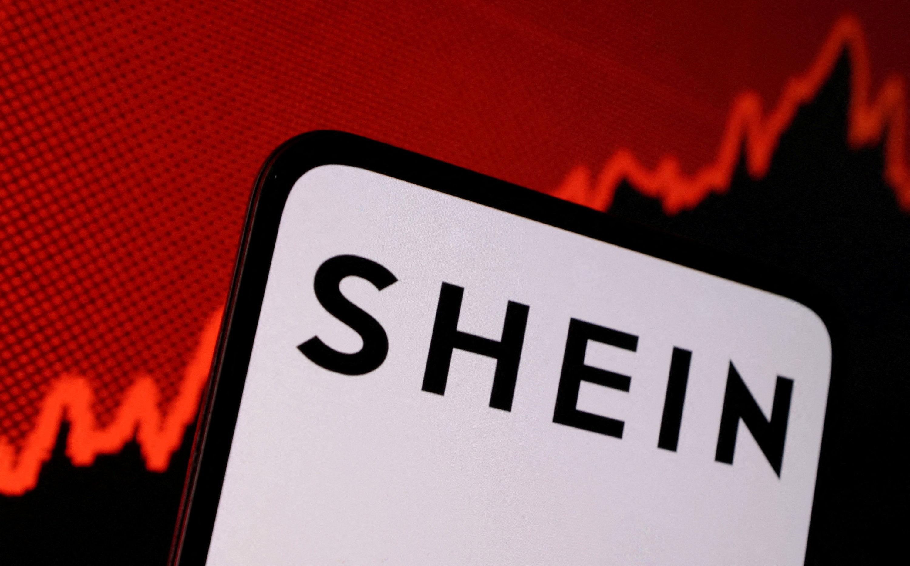 The image shows a smartphone with the word "SHEIN" on its screen, set against a background featuring a red line graph that appears to indicate a financial or market decline.