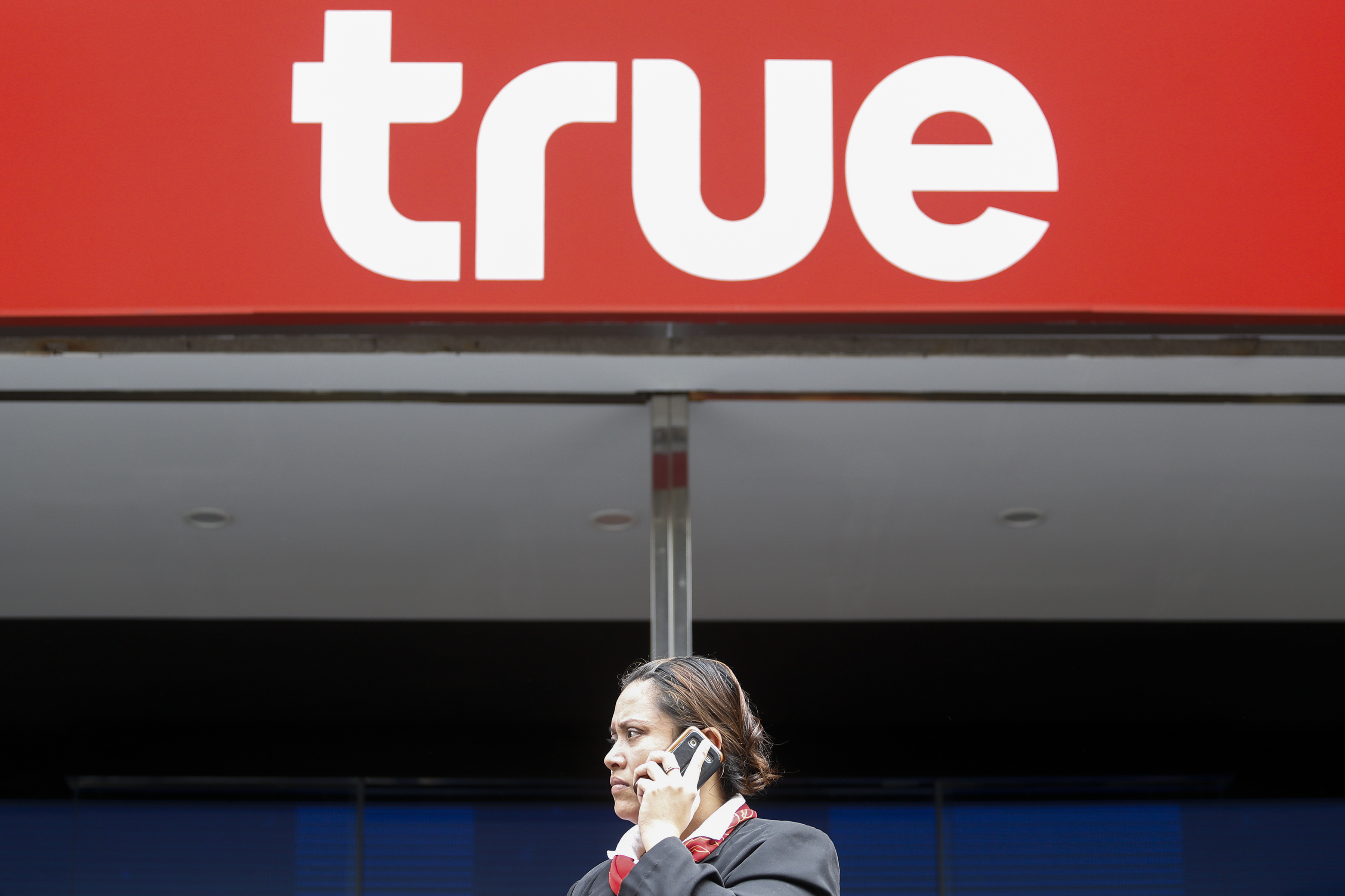 A woman speaks on a mobile phone as she stands next to the logo of True Corp in Bangkok