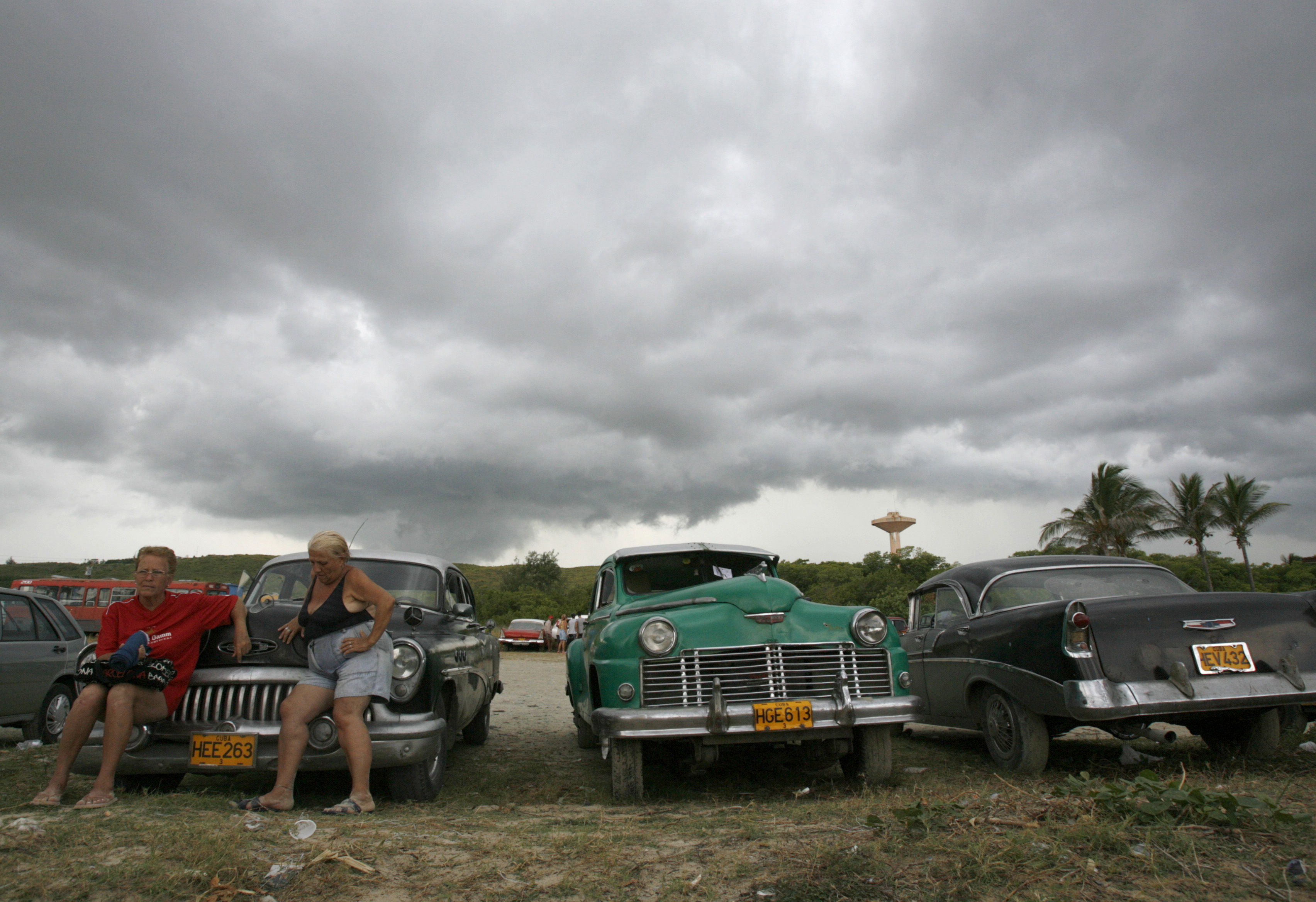 Women rest while sitting on a vintage car in a parking lot at the beach in Guanabo