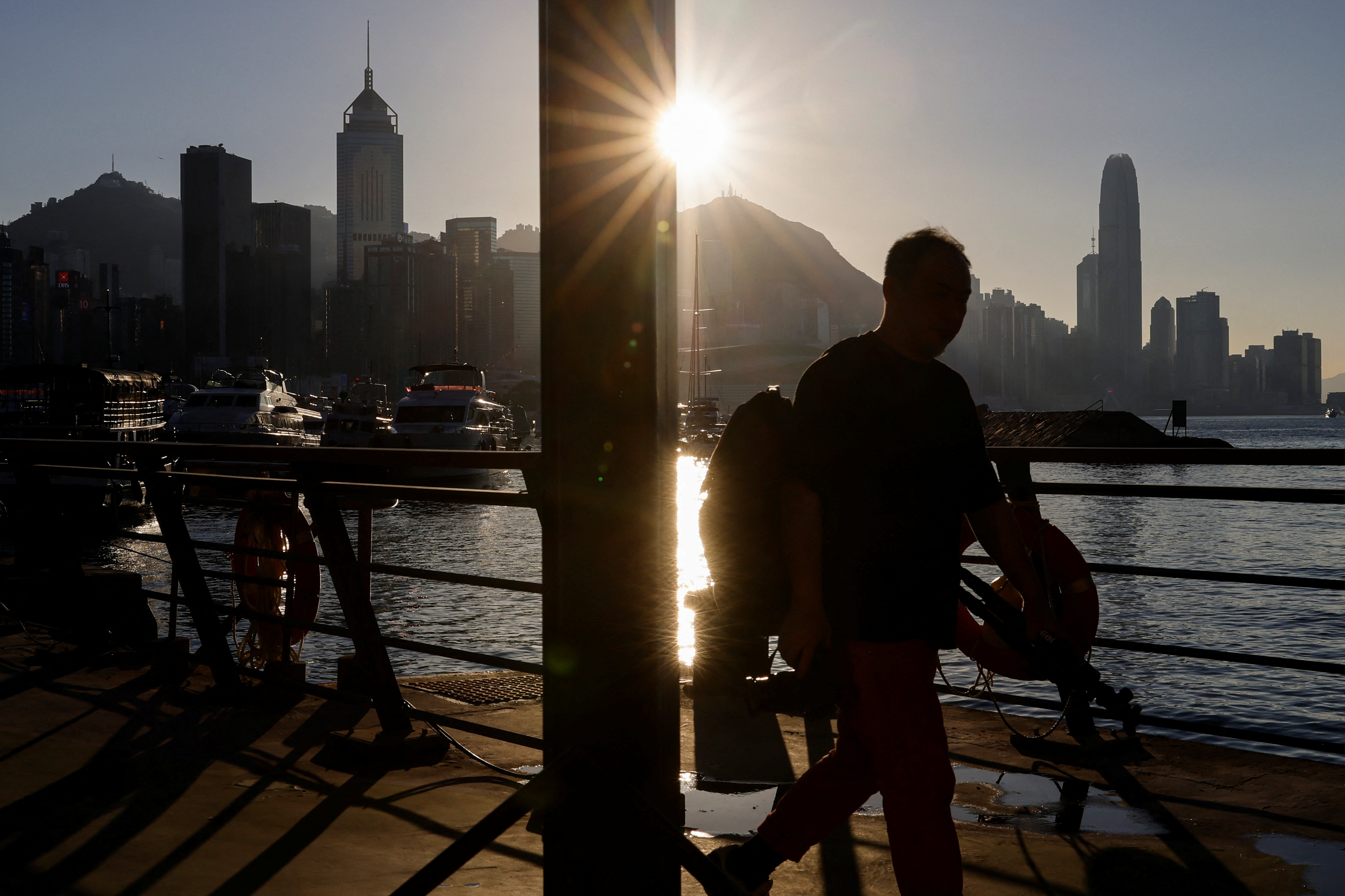 A man walks by the waterfront with skyline buildings across Victoria Harbor in Hong Kong