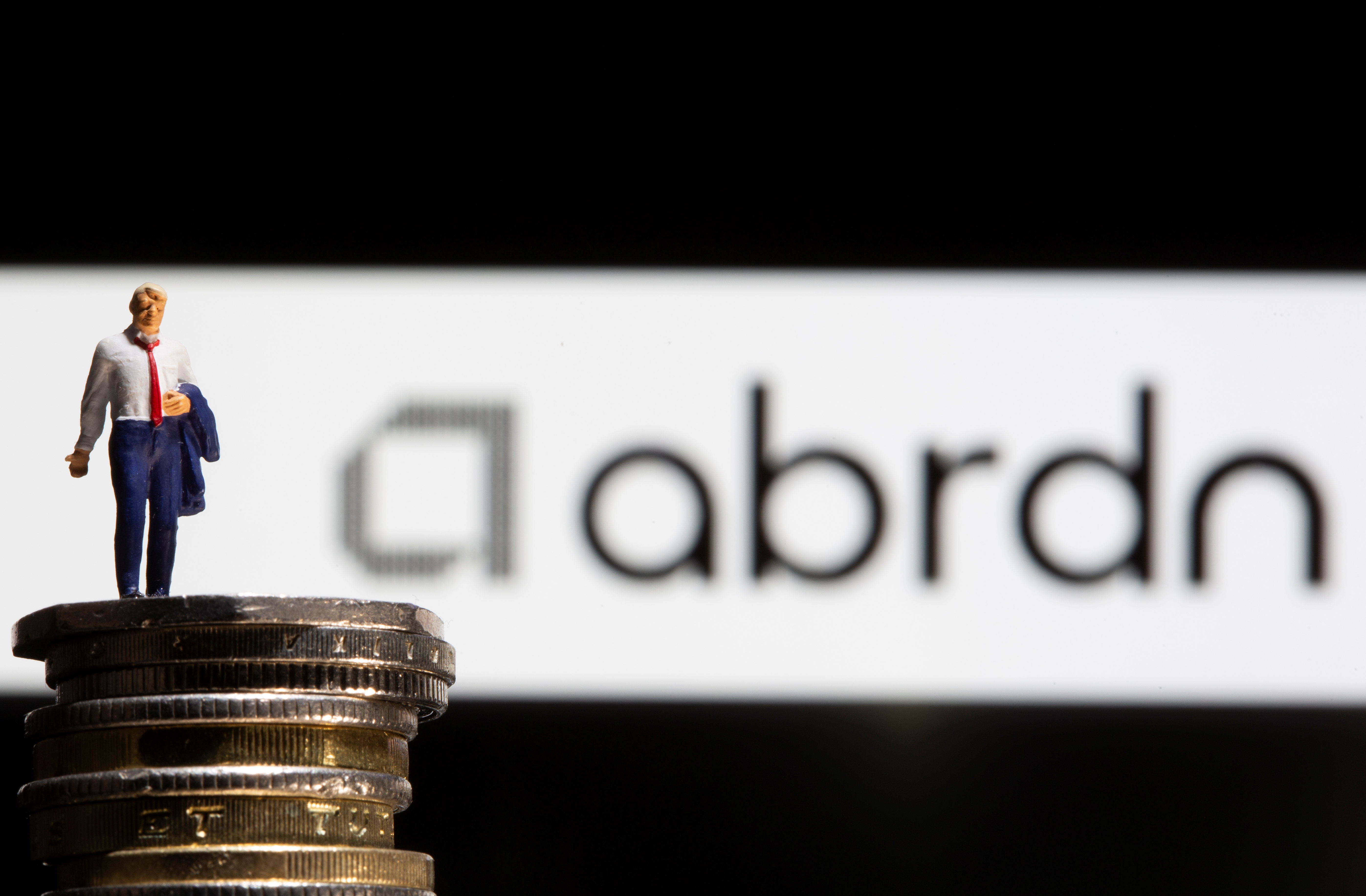 Businessman toy figure is placed on U.K. Pound coins in front of displayed Abrdn logo in this illustration taken