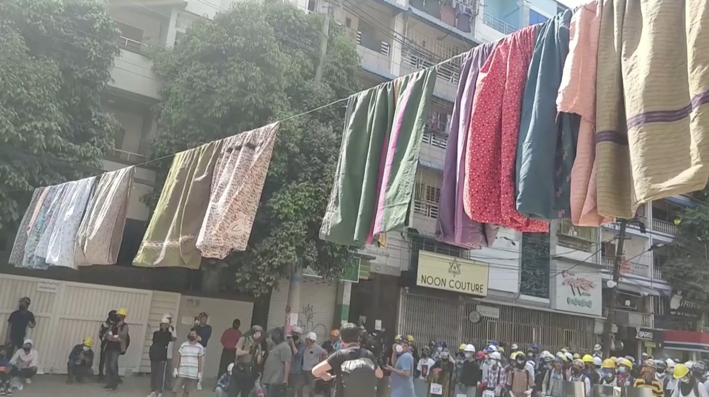 Traditional clothes hang on a rope as protesters hold shields in the background, in Yangon