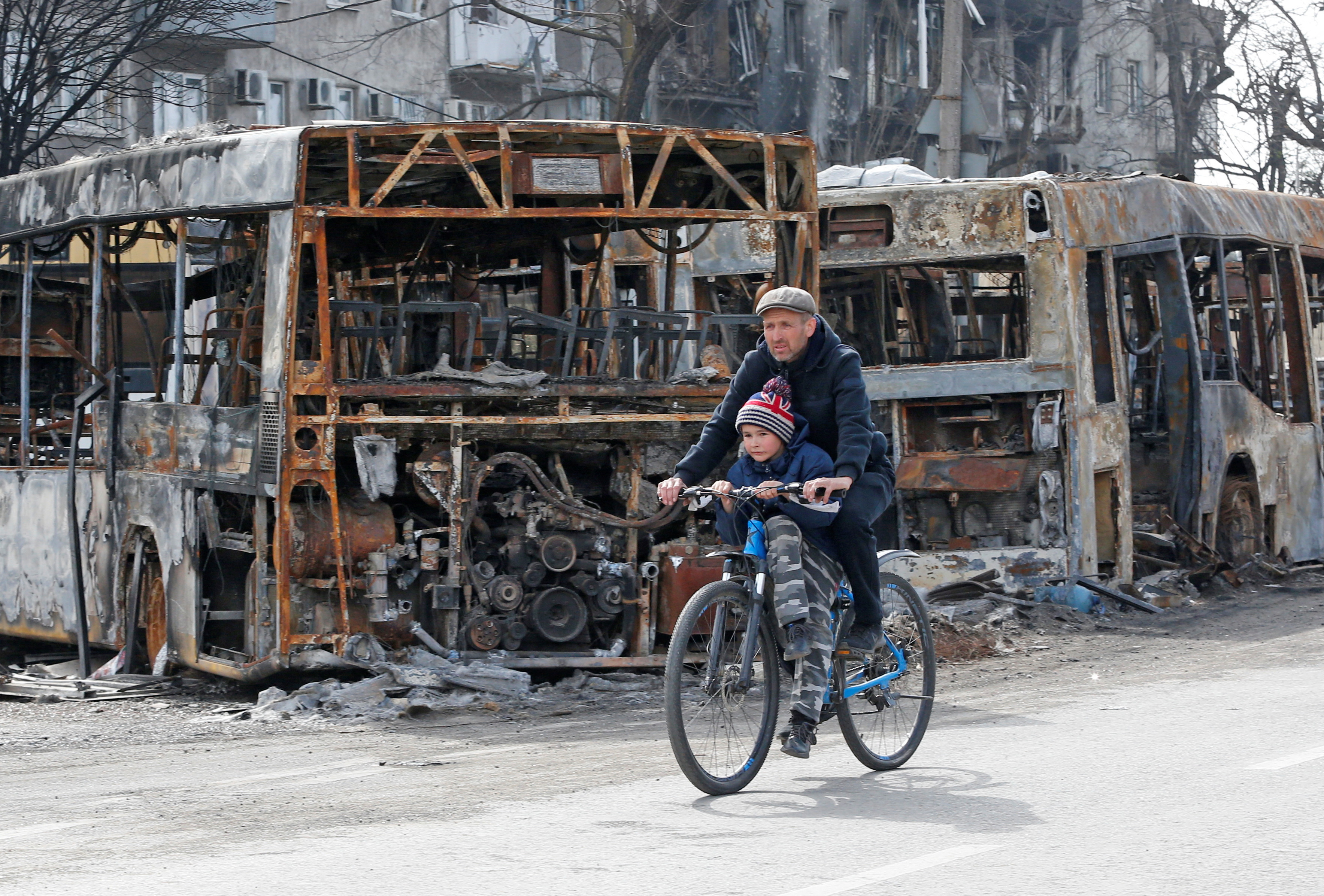 A man and a child ride a bicycle past burnt out buses in Mariupol