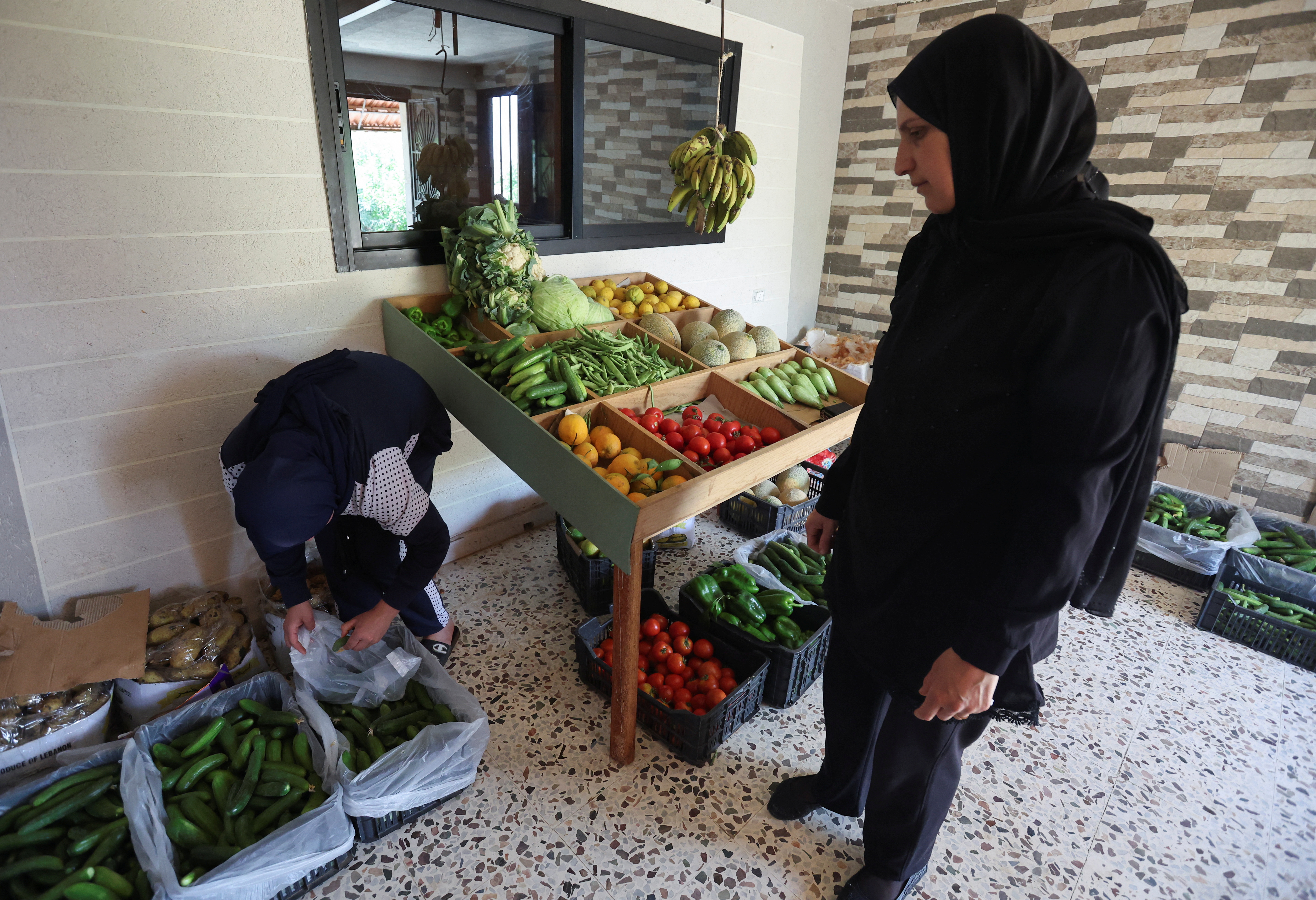 Khadija Shreim se encuentra cerca de su puesto de verduras y frutas mientras un cliente compra pepinos en la aldea de Houla