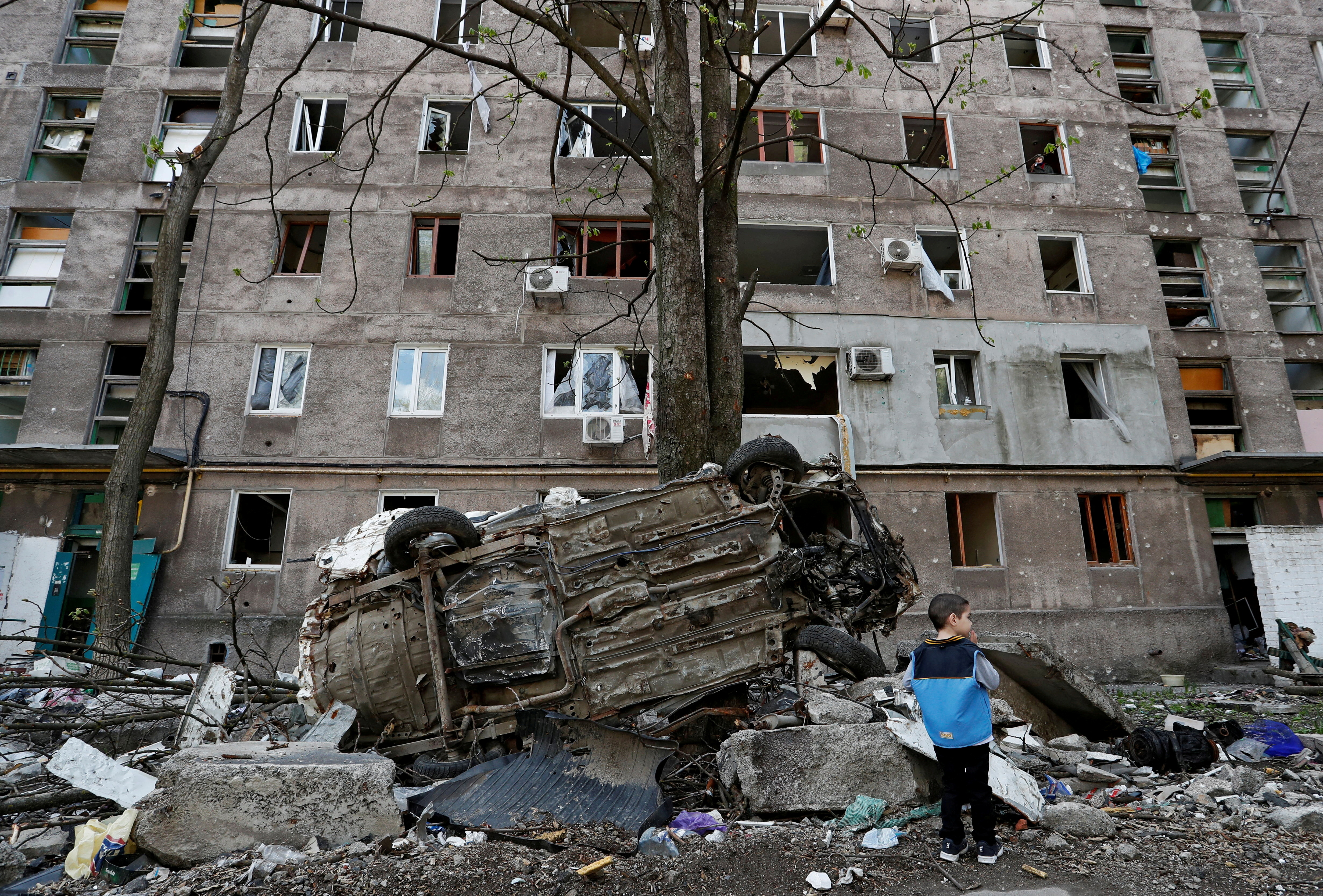 A boy stands next to a wrecked vehicle in front of a damaged apartment building in Mariupol