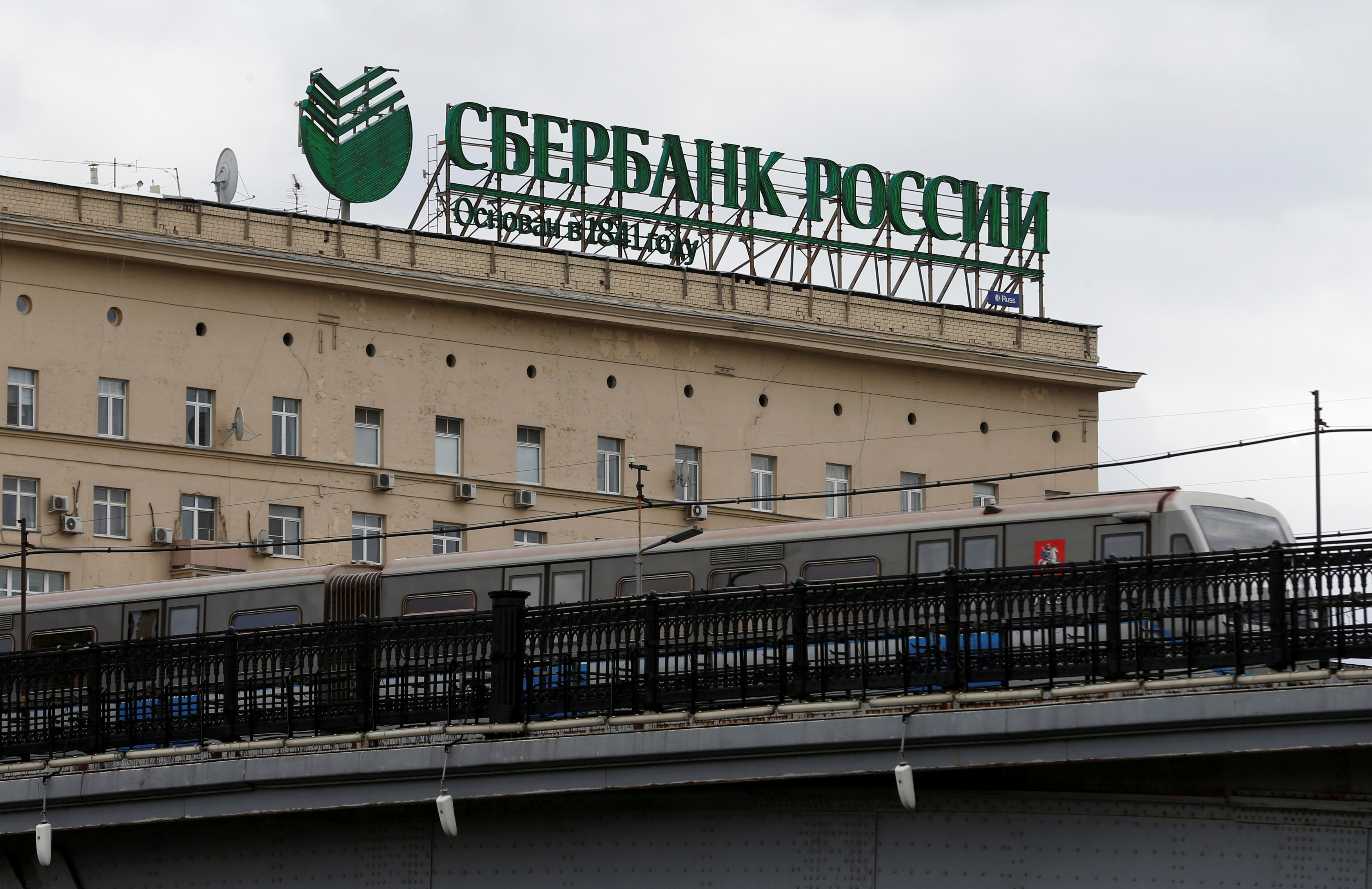 Metro train moves on bridge with logo of Sberbank on top of building seen in background in Moscow