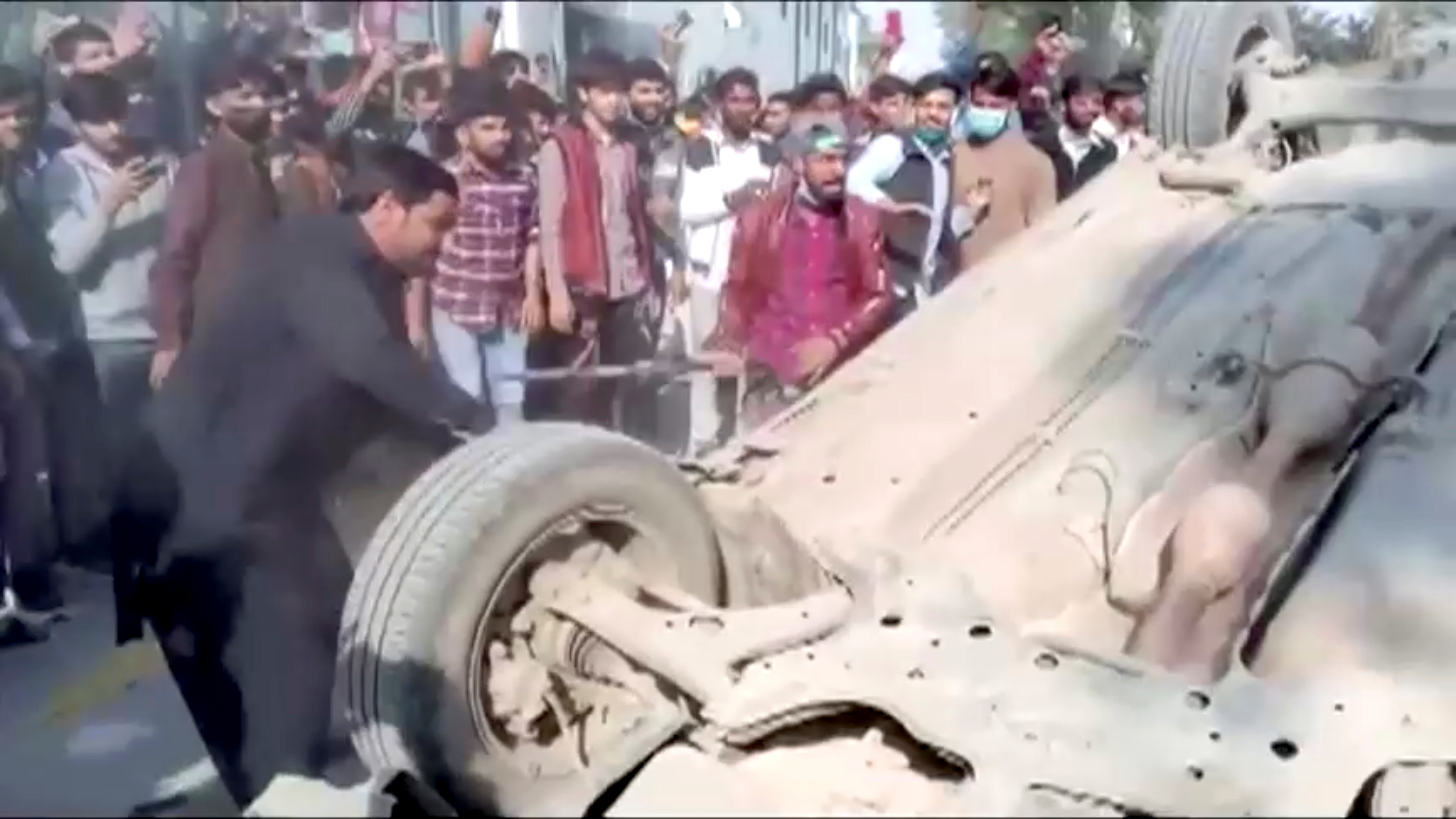 A mob hits an overturned car with sticks after an attack on a factory in Sialkot