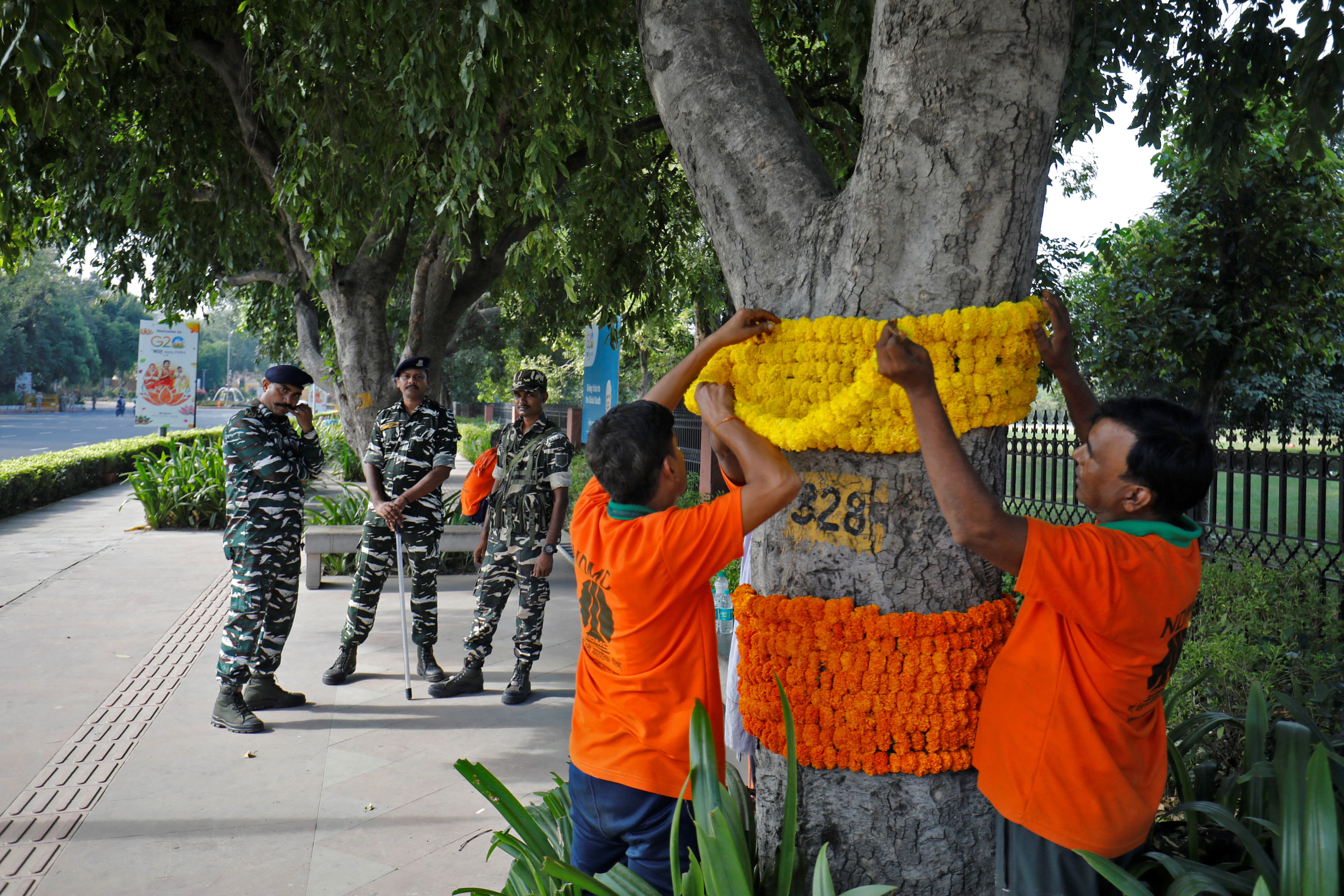 Men decorate a tree with flower garlands as security force personnel stand guard on a pavement ahead of the G20 Summit in New Delhi