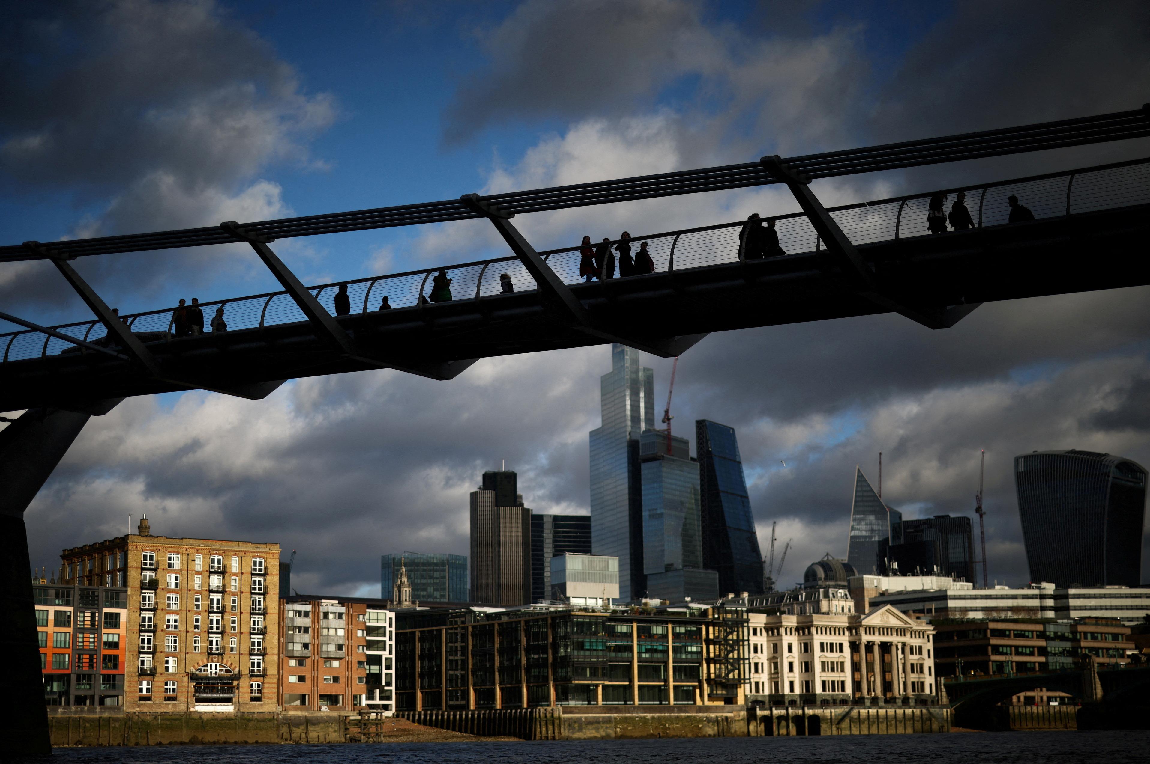 People walk over the Millennium Bridge with the City of London financial district in the background, in London