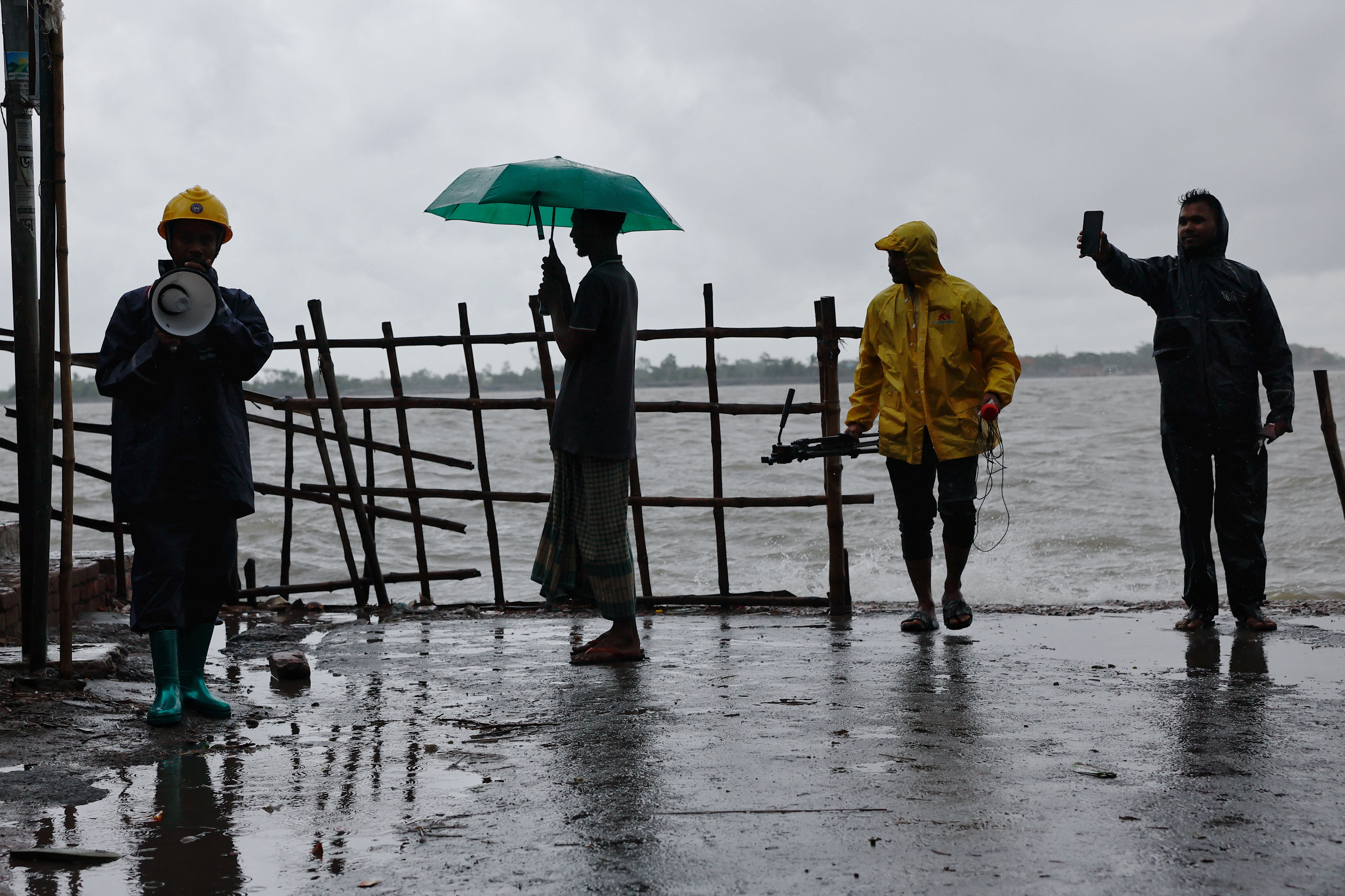 People stand on the bank of Kholpetua river while a volunteer alerts people before the Cyclone Remal hits the country in the Shyamnagar area of Satkhira