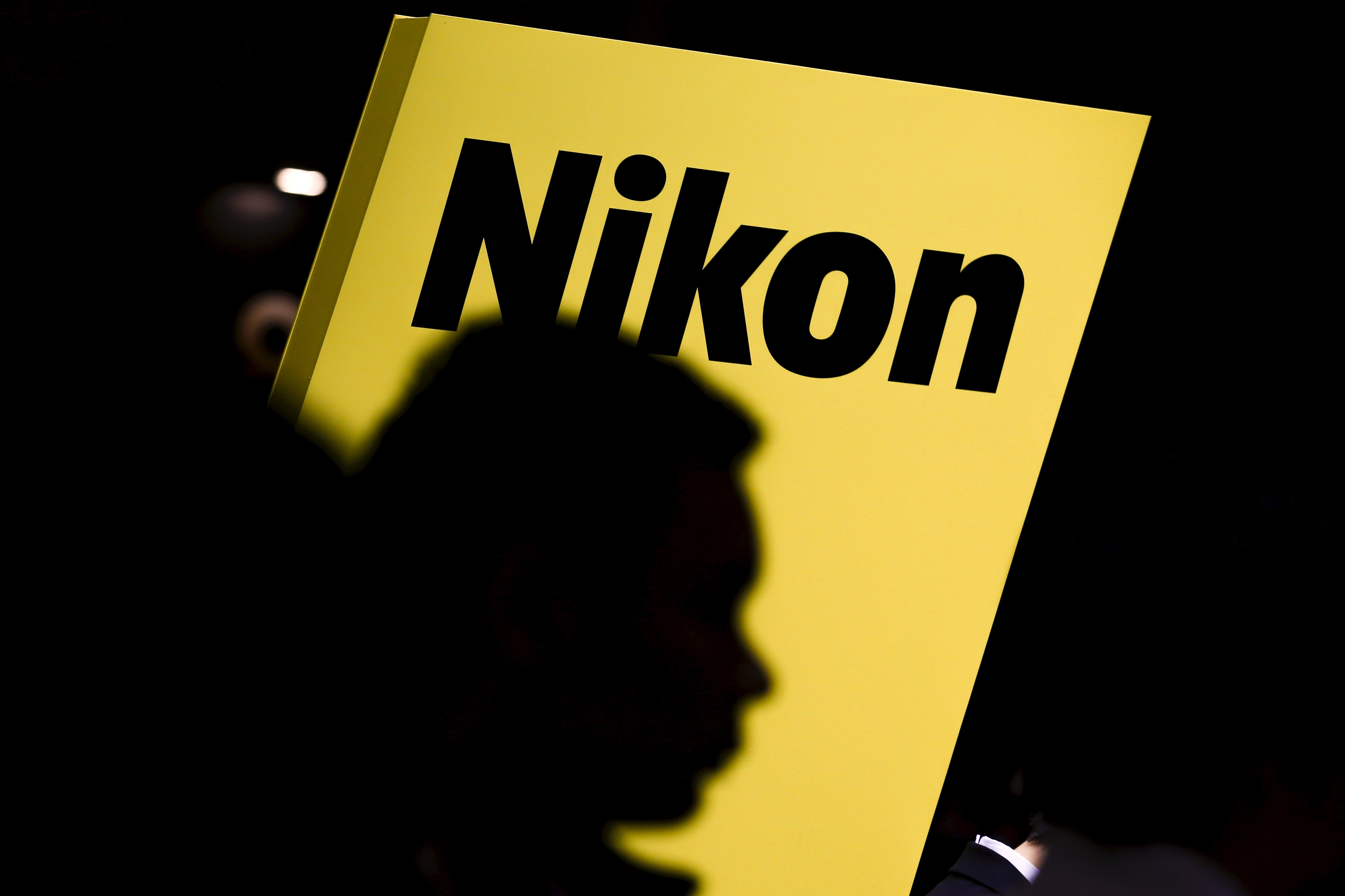 People are silhouetted against a display of the Nikon brand logo at the CP+ camera and photo trade fair in Yokohama