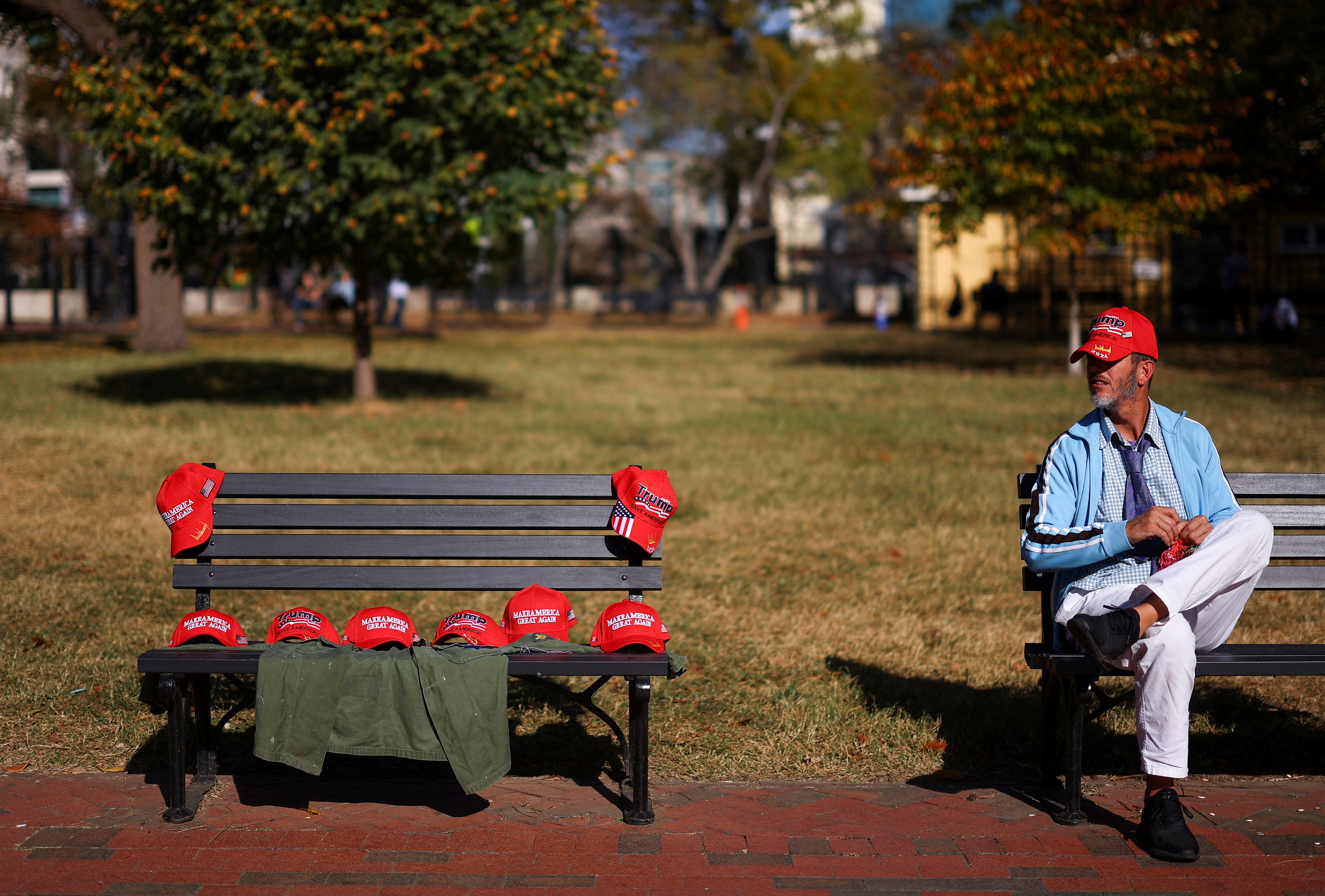 A man sits next to a bench with MAGA hats, outside the White House