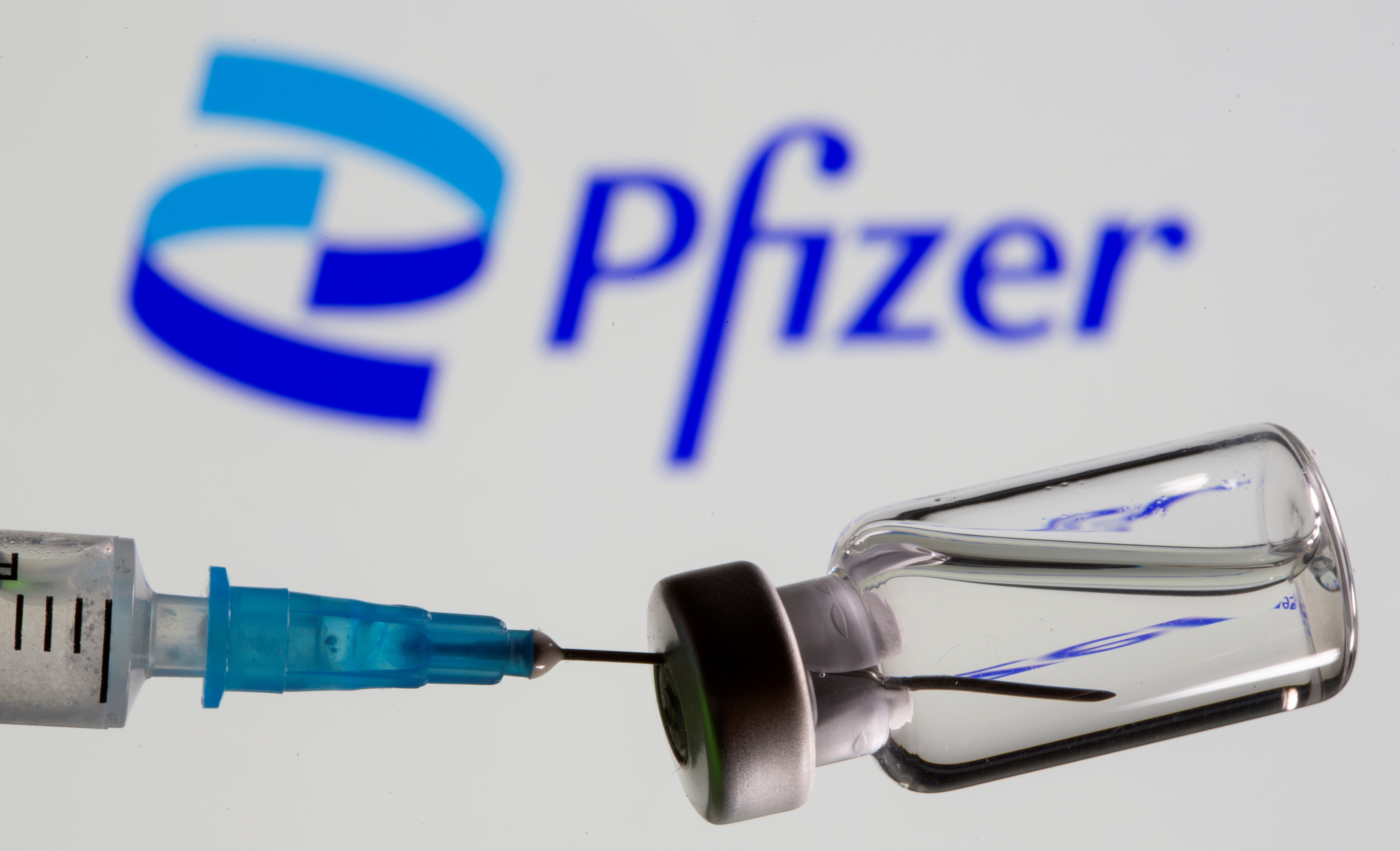 Syringe and vial are seen in front of displayed new Pfizer logo in this illustration taken