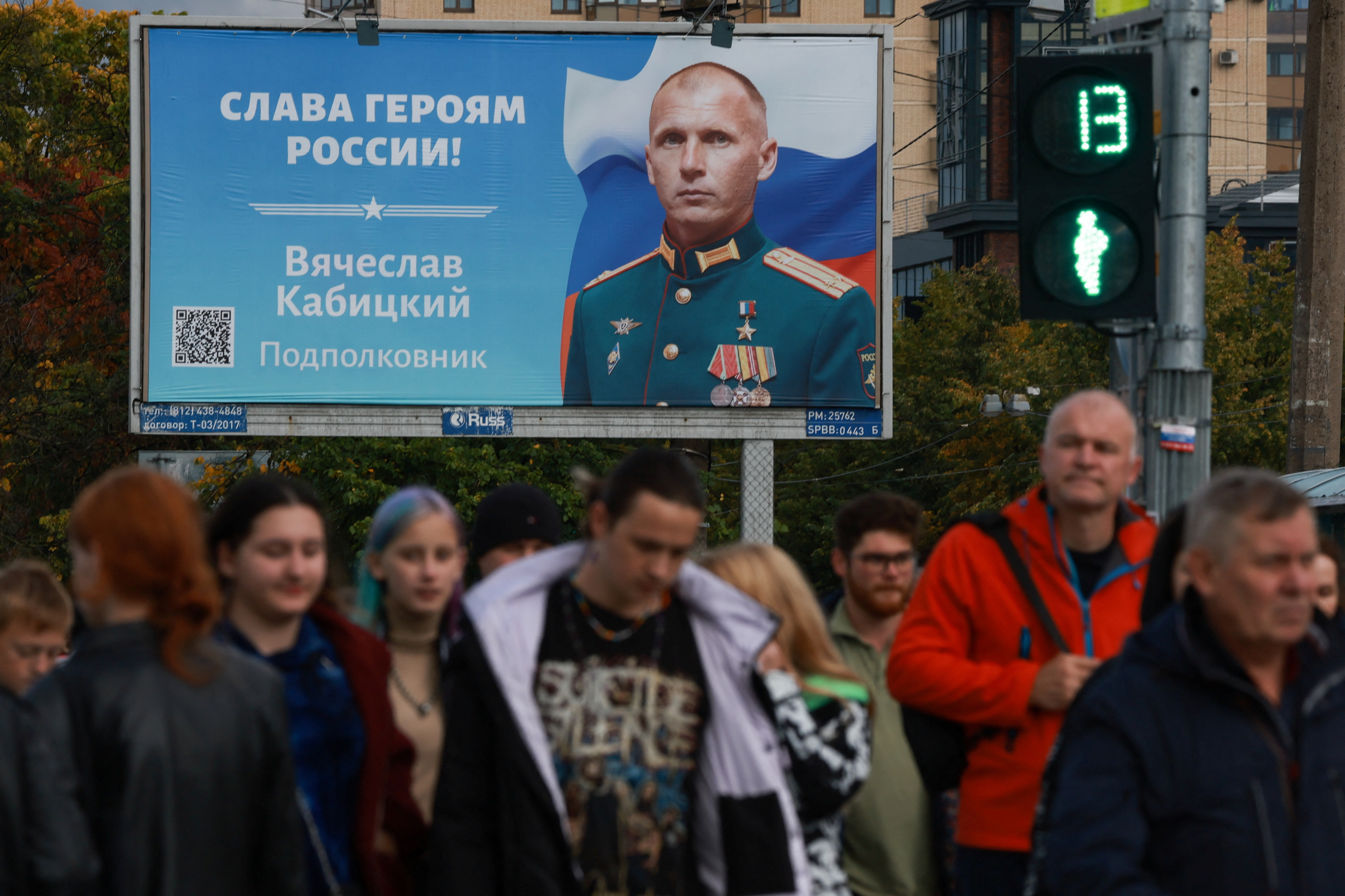 Pedestrians cross a street near a board with a portrait of a Russian service member in Saint Petersburg