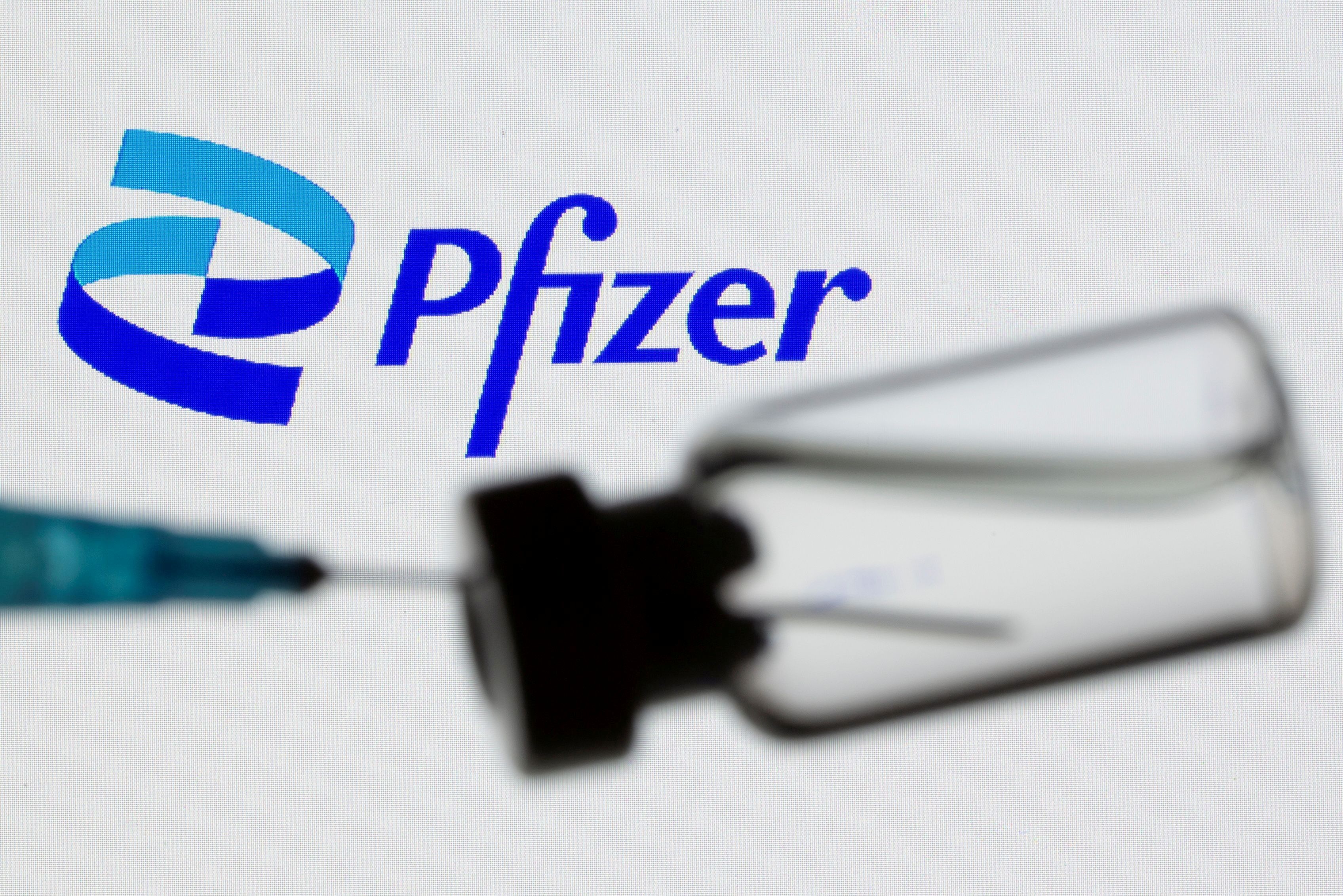 A syringe and vial are seen in front of a displayed Pfizer logo in this illustration