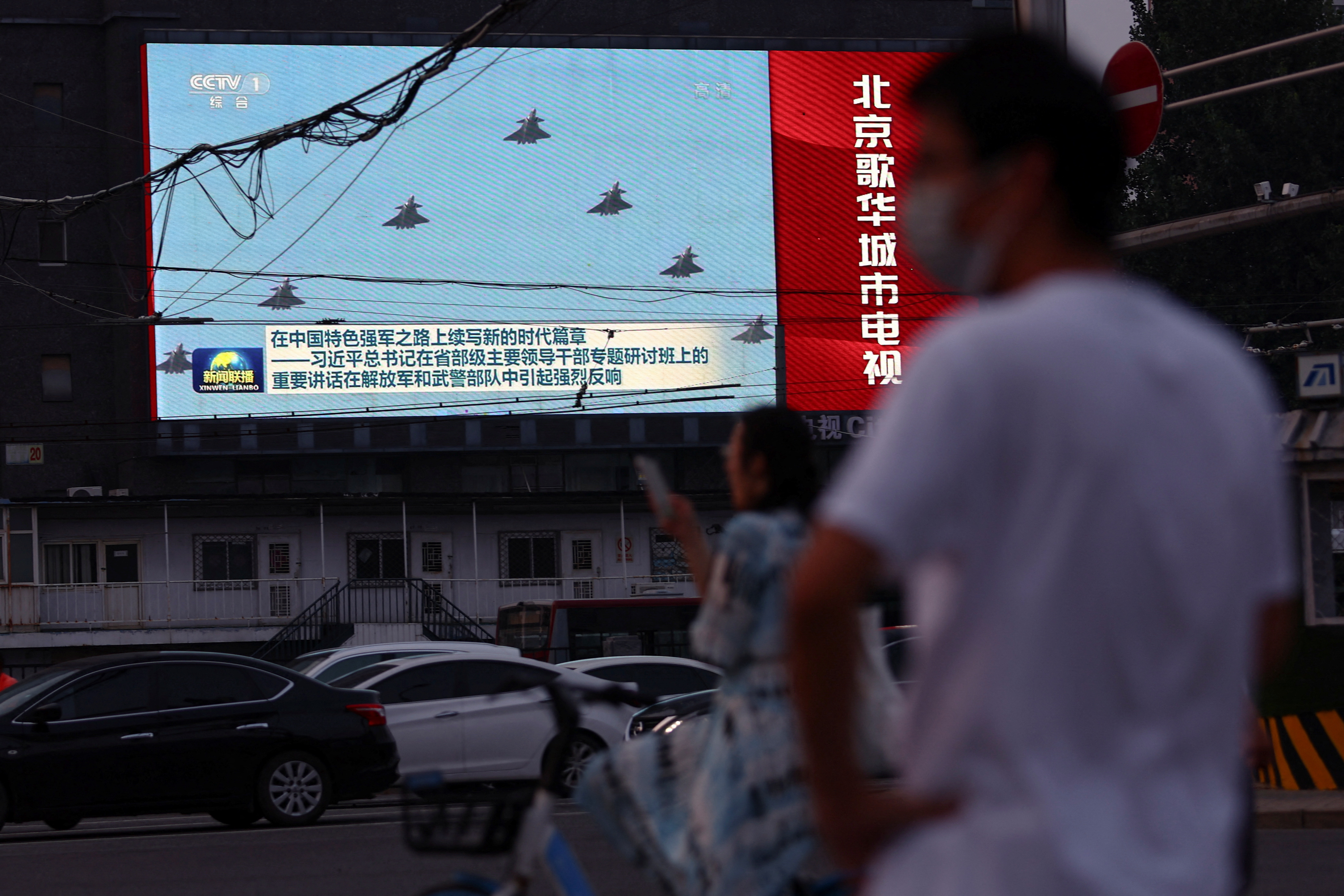 Pedestrians wait at an intersection near a screen showing footage of Chinese People's Liberation Army (PLA) aircraft during an evening news programme, in Beijing