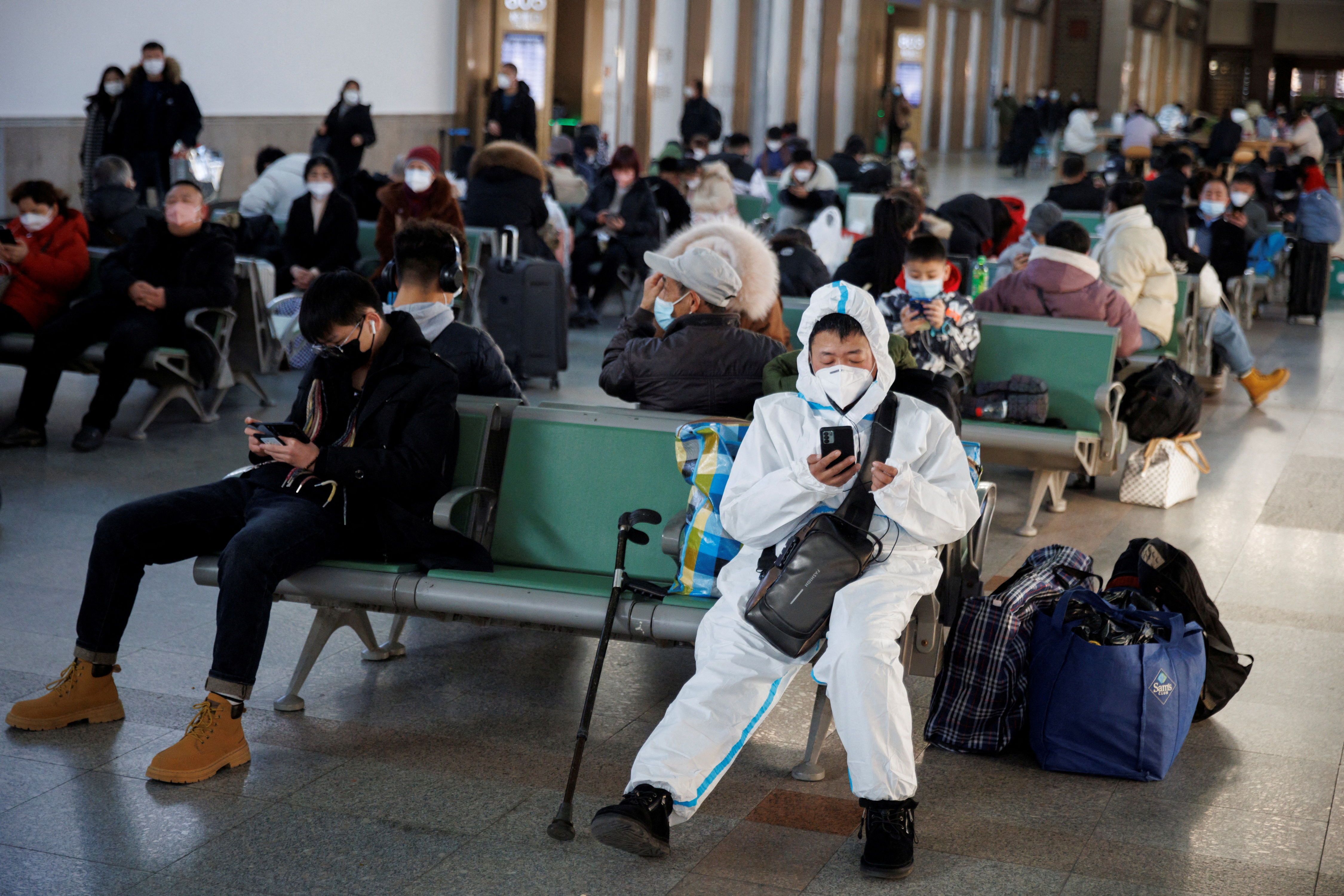 A person wearing a protective suit sits in Beijing Railway Station as passengers wait to board a train to travel for Spring Festival ahead of Chinese Lunar New Year festivities in Beijing