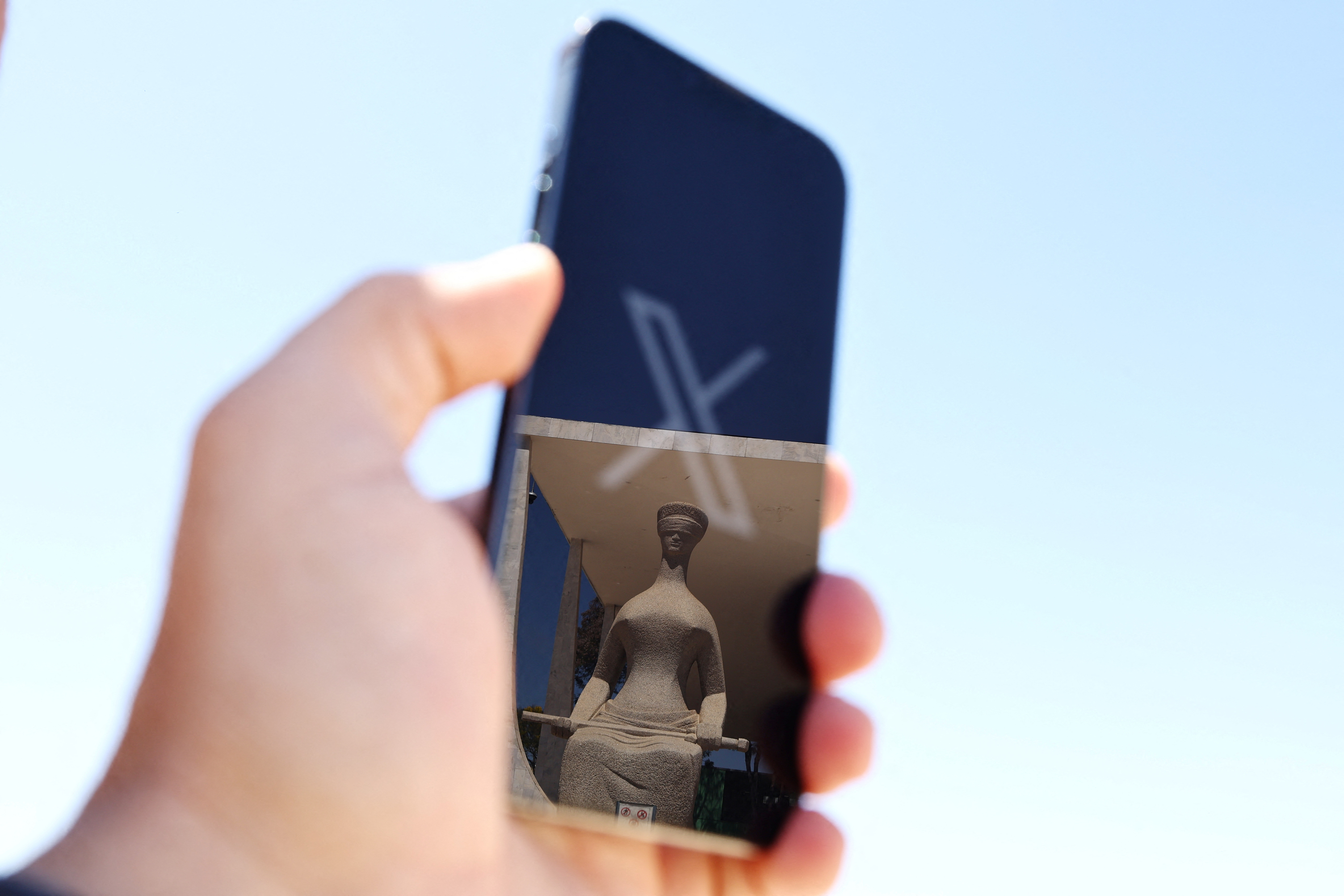 The logo of social media platform X is seen on a mobile phone next to a reflection of Alfredo Ceschiatti's sculpture The Justice, outside Brazil's Supreme Court in this illustration taken August 30, 2024, in Brasilia