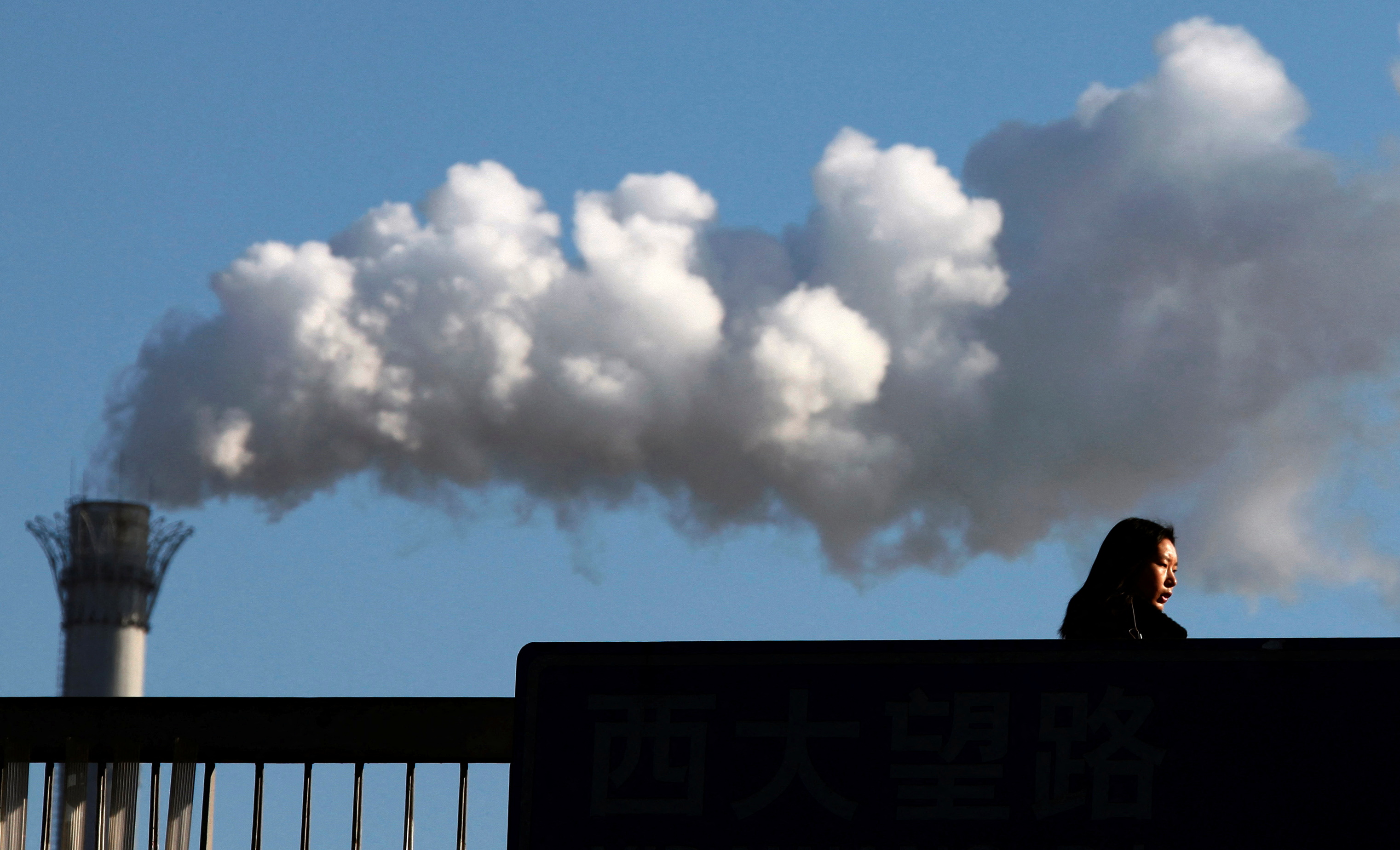A woman walks across a bridge in front of a chimney billowing smoke from a coal-burning power station in central Beijing.