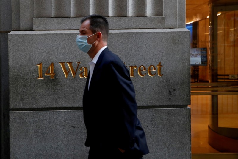 A man wearing a protective face mask walks by 14 Wall Street in the financial district of New York