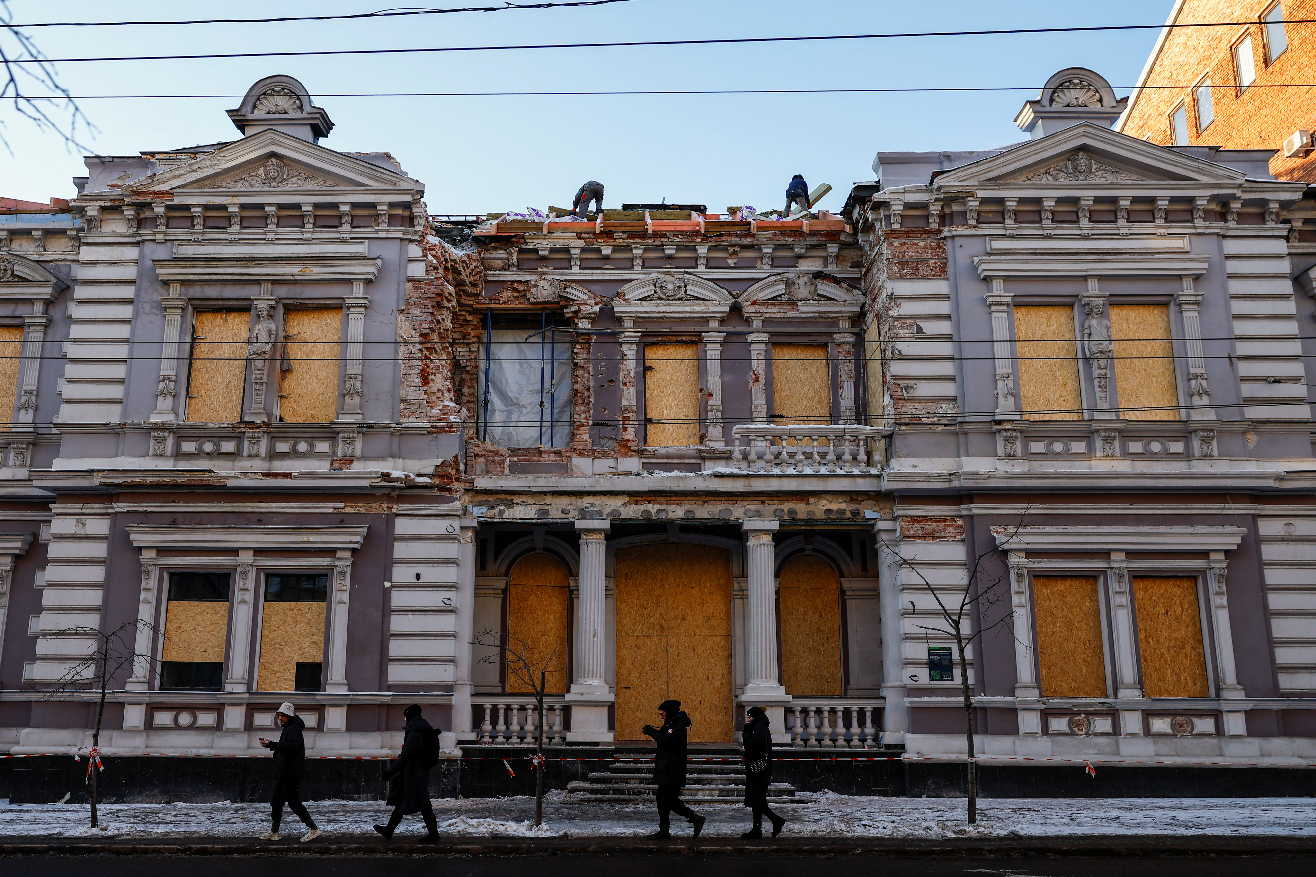 People walk past as workers repair the roof of a historical building, damaged during one of the latest Russian drone strikes, in Kharkiv