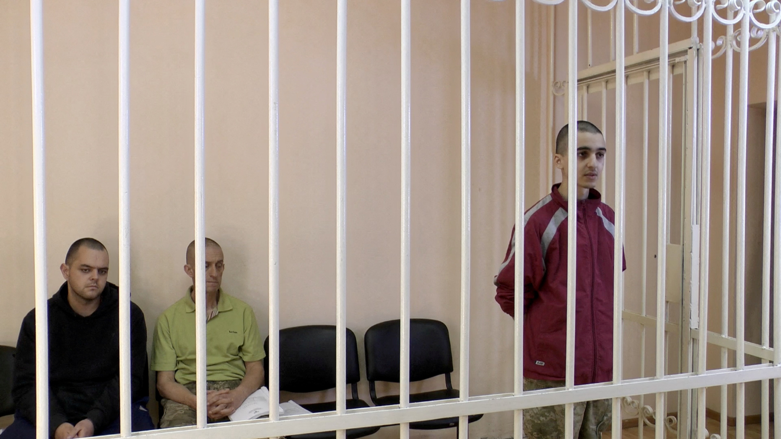 Still image showing Britons Aiden Aslin and Sean Penner and Moroccan Ibrahim Saadoun in a cage in a courtroom in a place known as Donetsk.