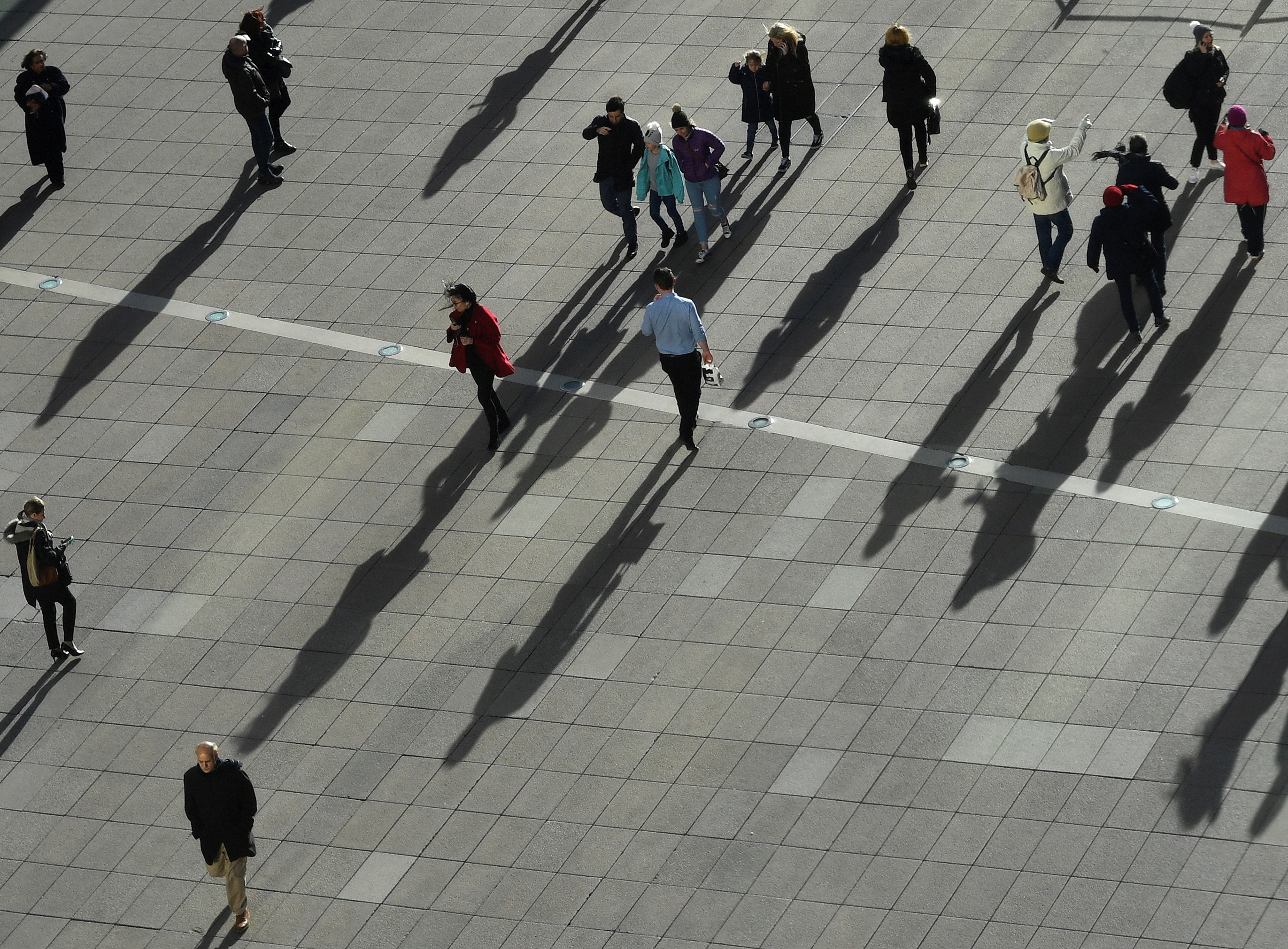 People cast long shadows in the winter sunlight as they walk across a plaza in the Canary Wharf financial district of London