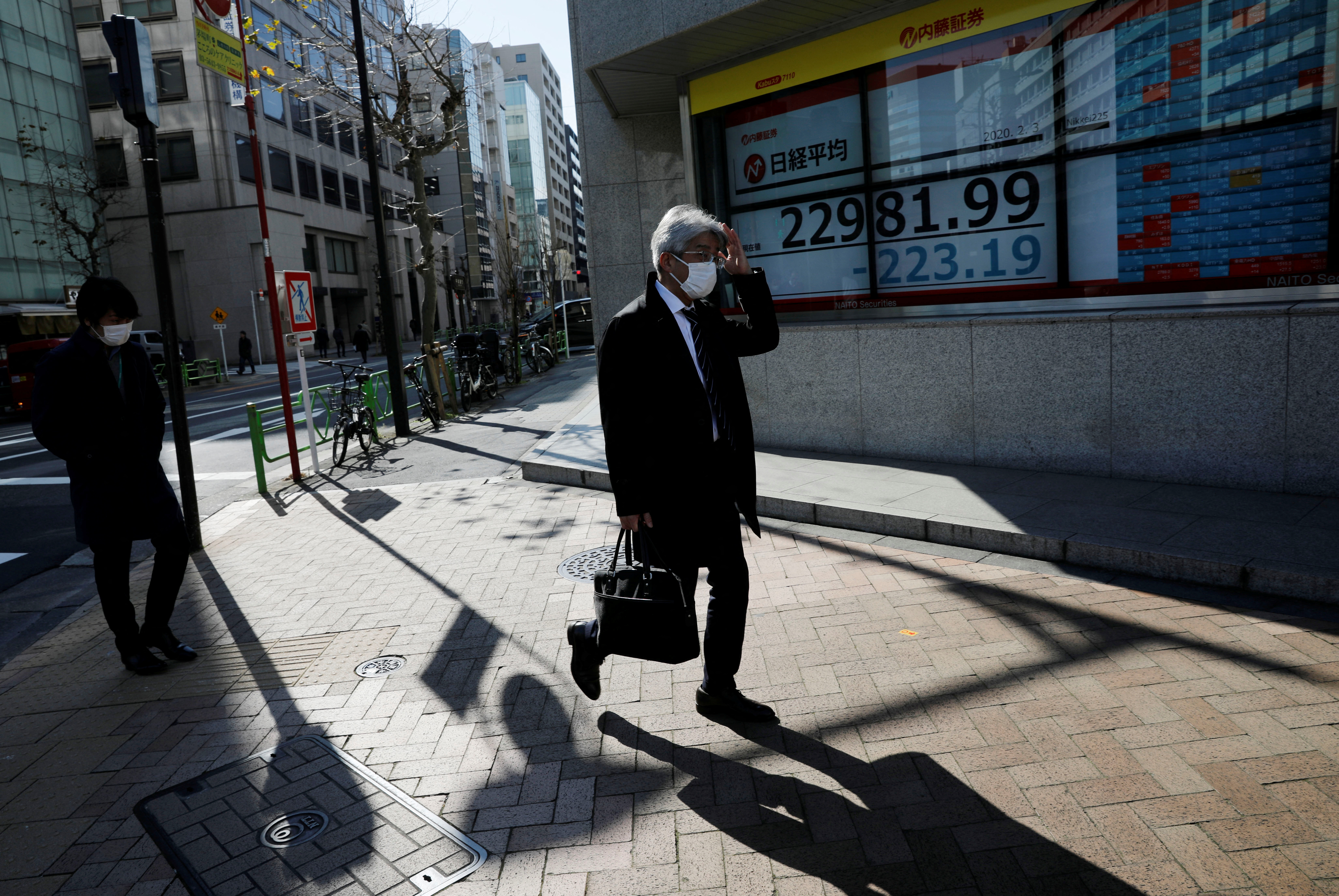 People wearing surgical masks walk past a screen showing Nikkei index outside a brokerage in Tokyo