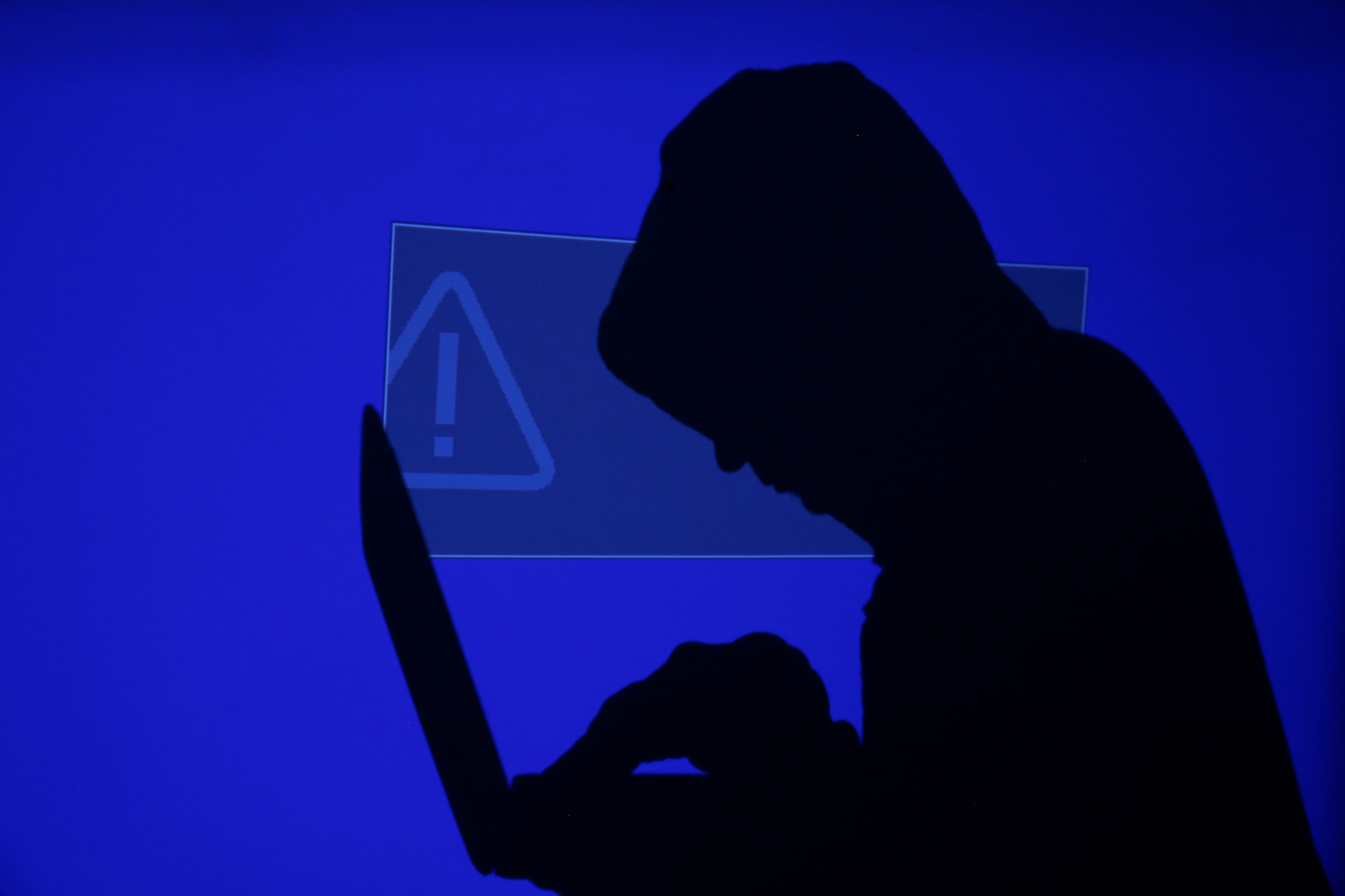 Hooded man holds laptop computer as blue screen with an exclamation mark is projected on him in this illustration picture