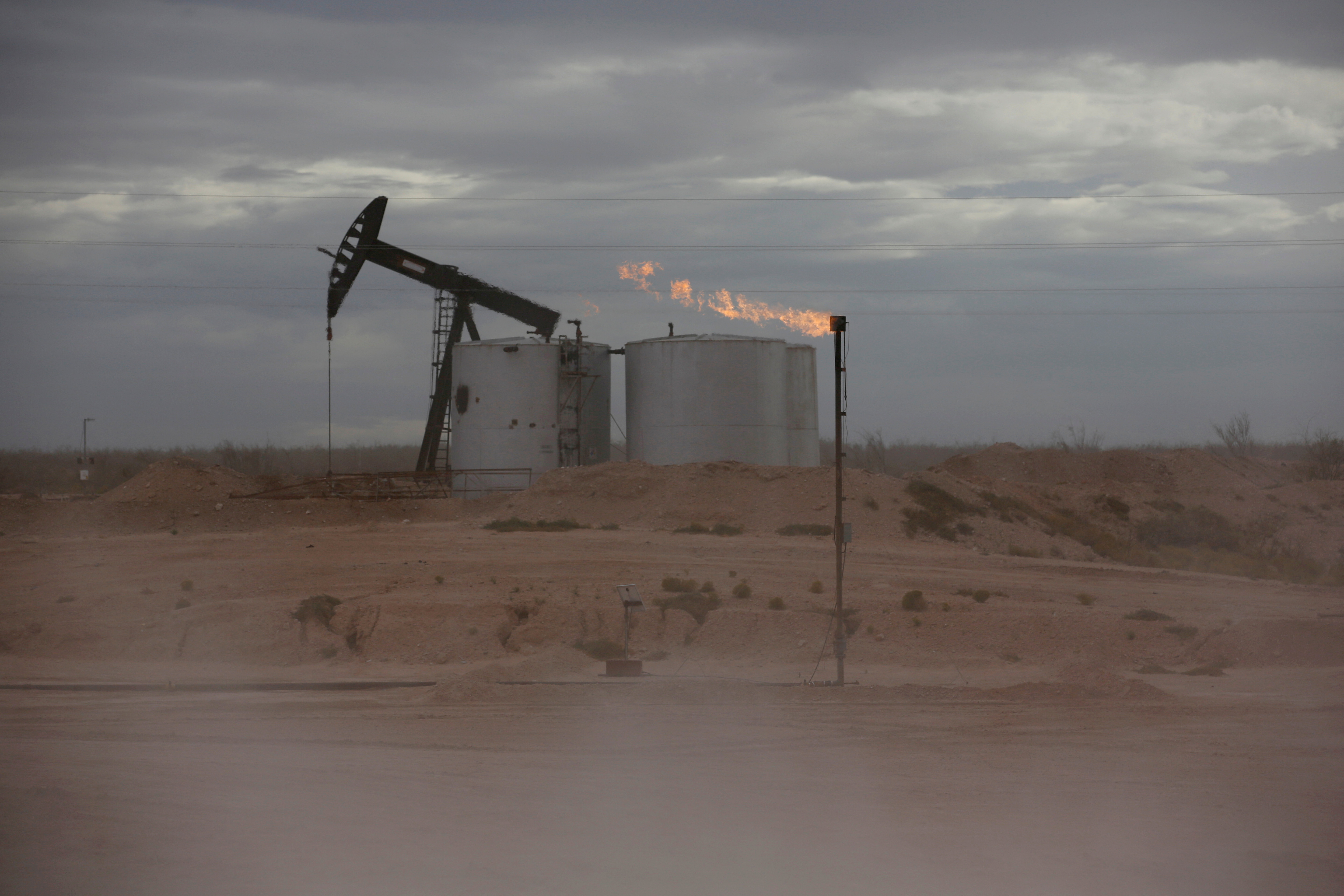 Dust blows around a crude oil pump jack and flare burning excess gas at a drill pad in the Permian Basin in Loving County