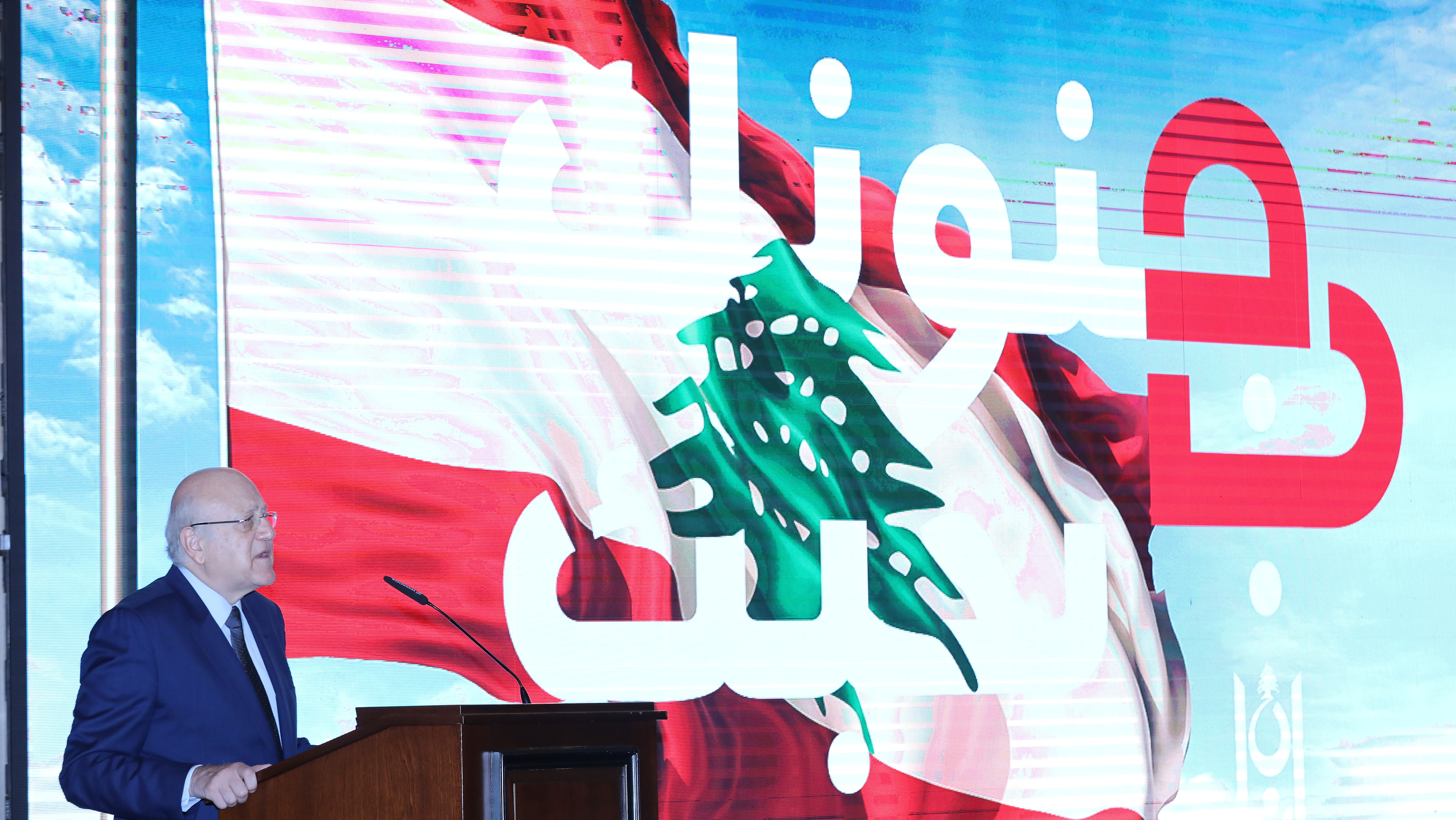 Lebanese Prime Minister Najib Mikati speaks as he stands near a slogan that reads 'We love you in your madness' displayed on a screen at the government palace in Beirut