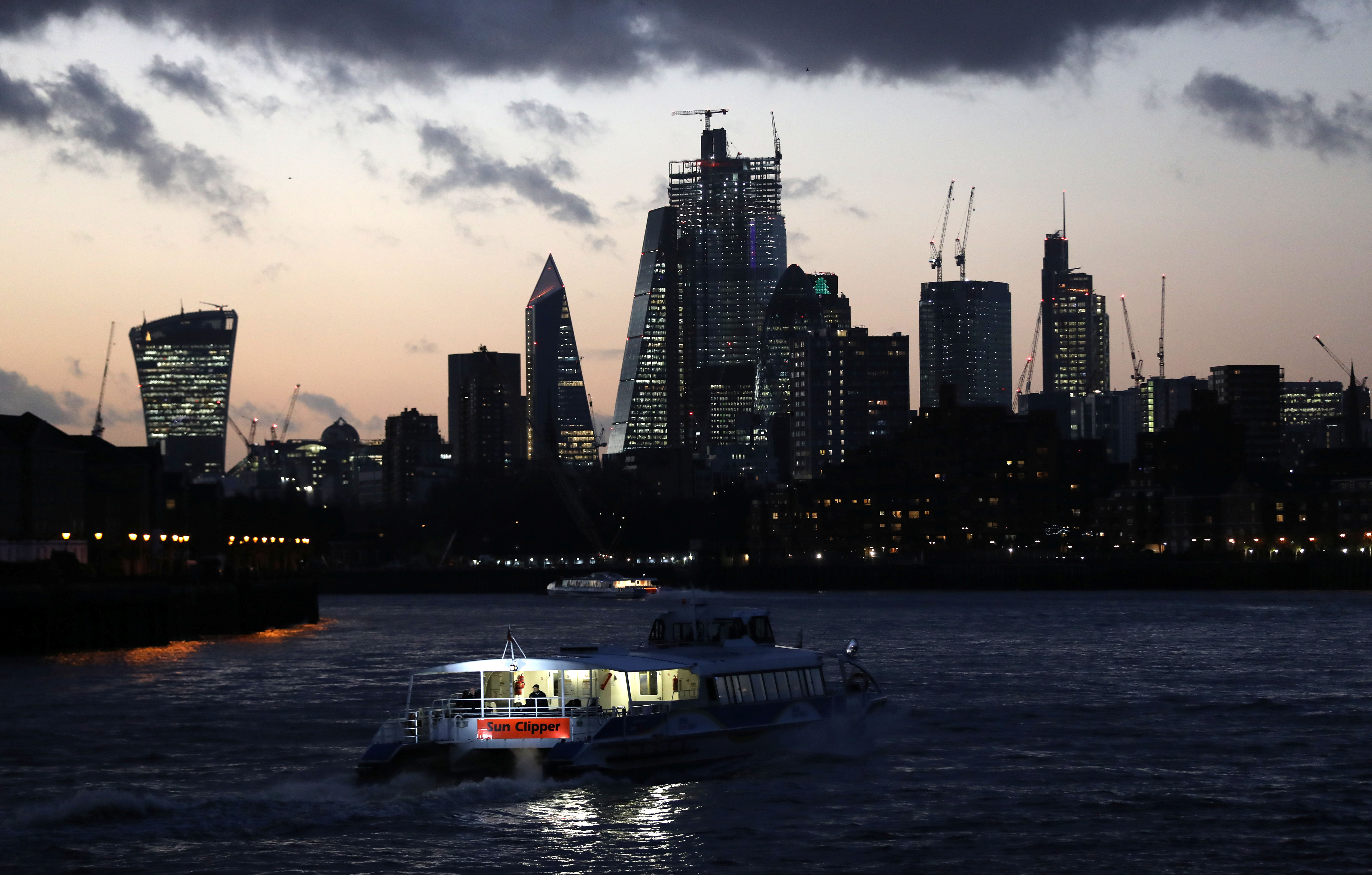 A river boat cruises down the River Thames as the sun sets behind the Canary Wharf financial district of London