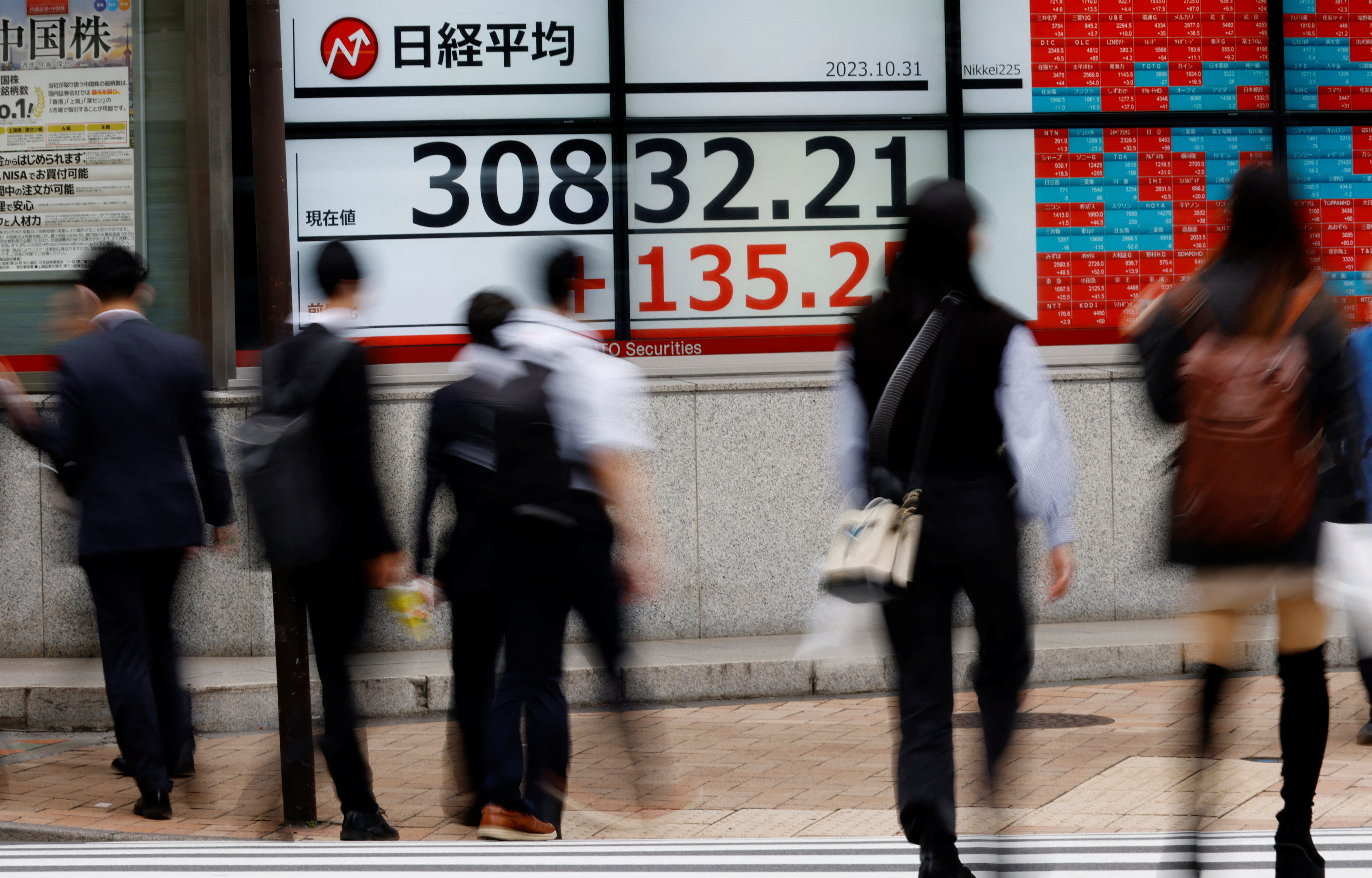 Pedestrians walk past an electronic board displaying the Nikkei stock average, outside a brokerage firm in Tokyo
