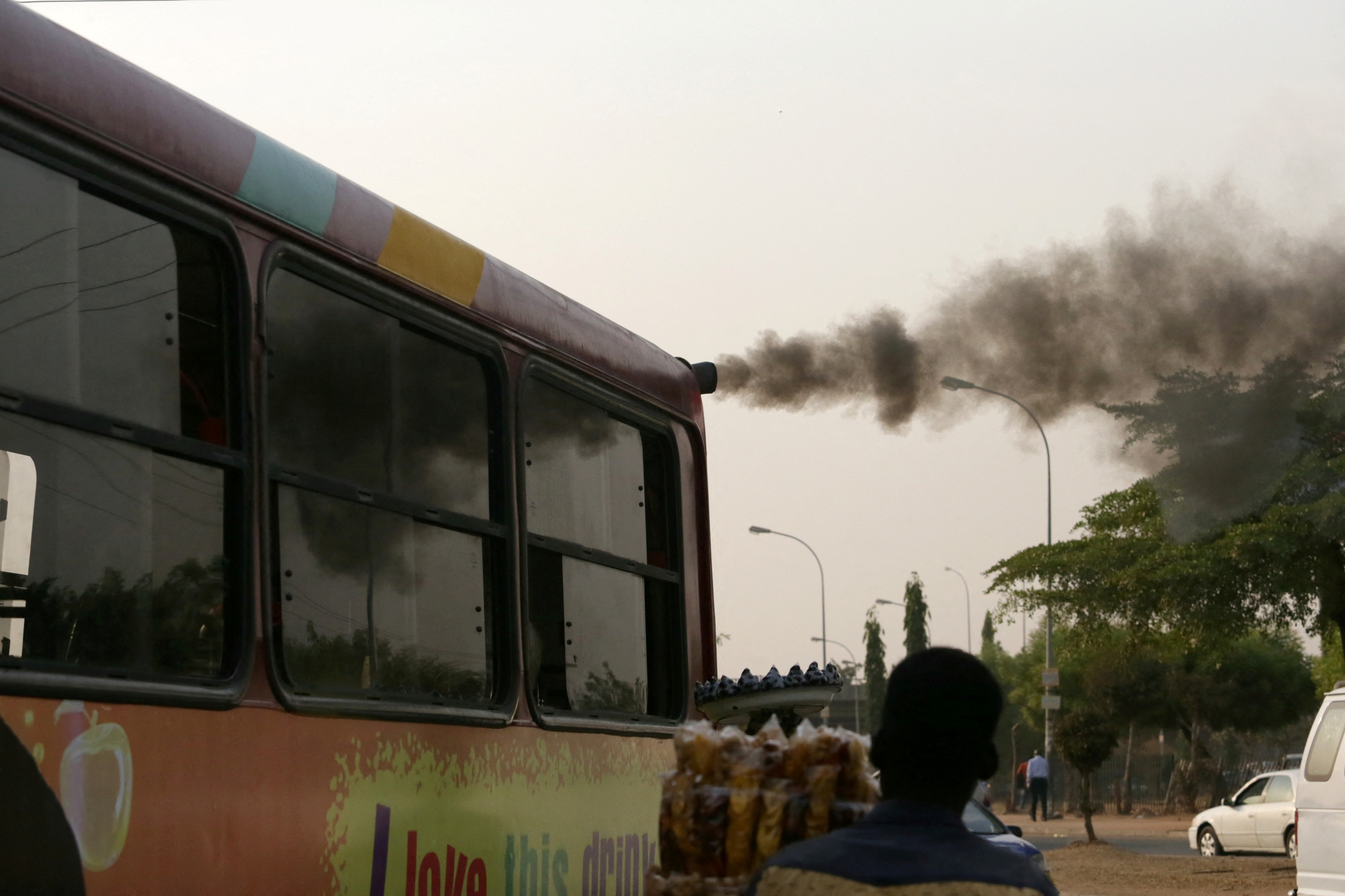 A man sells plantain chips near a bus with smoke seen from its exhaust at a bus park in Abuja