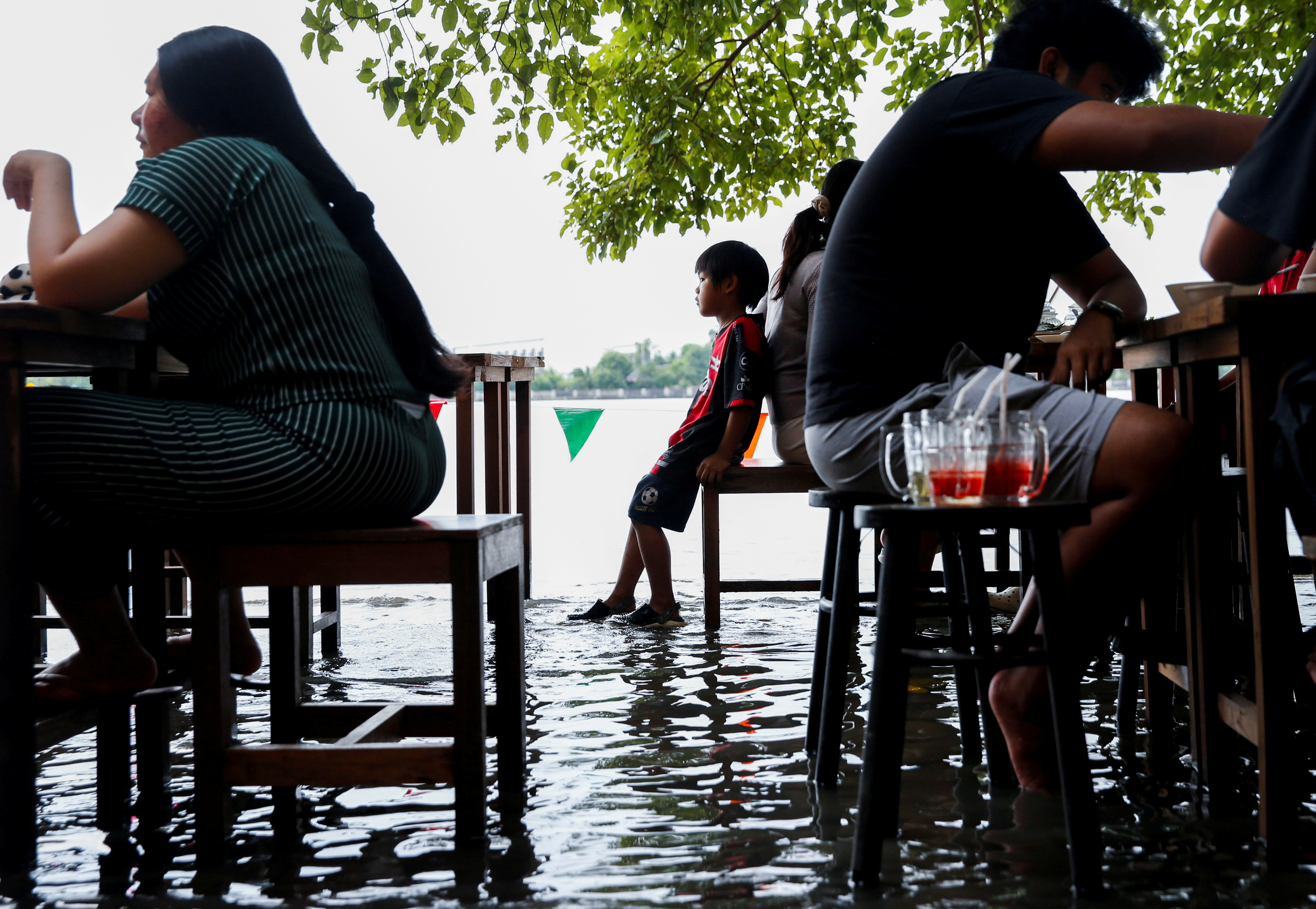 People eat food at a flooded restaurant, where patrons stand up from their tables every time the waves come in, on a river bank in Nonthaburi