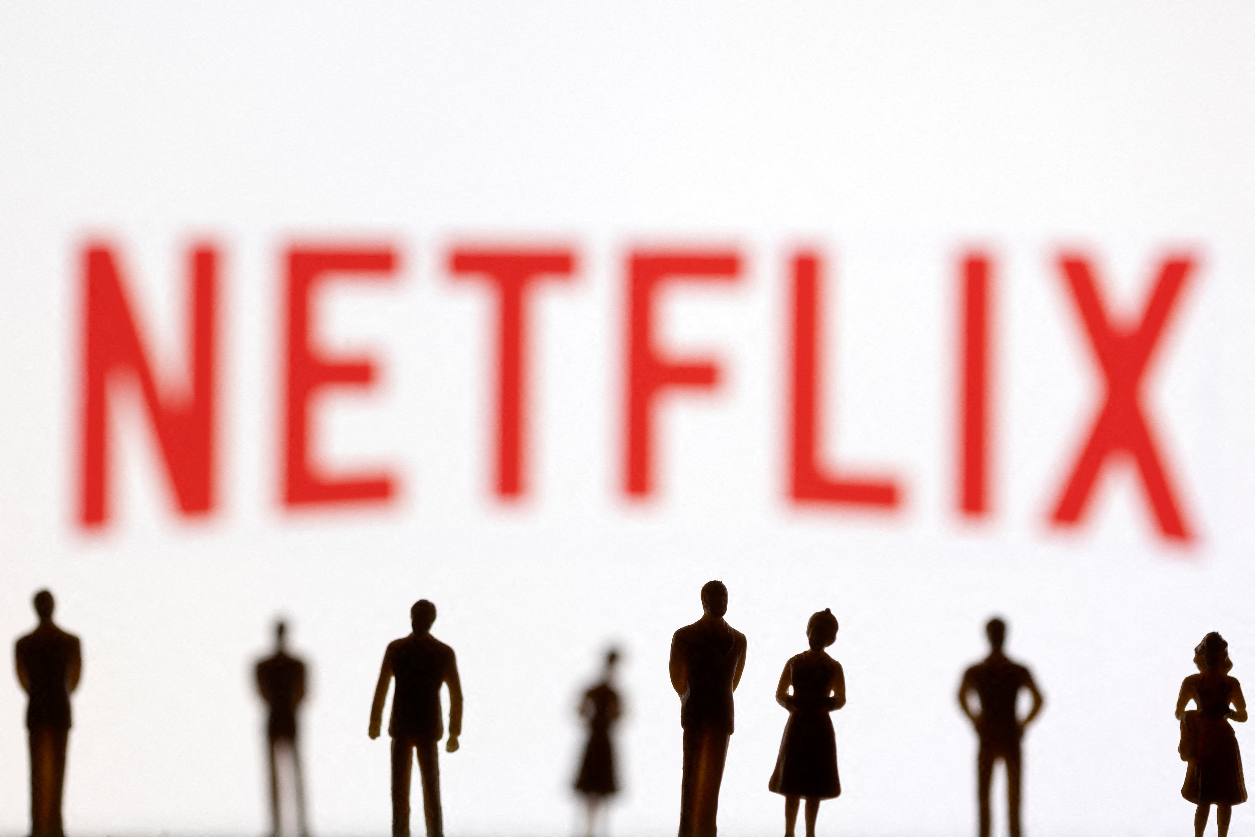 Toy figures of people are seen in front of the displayed Netflix logo, in this illustration