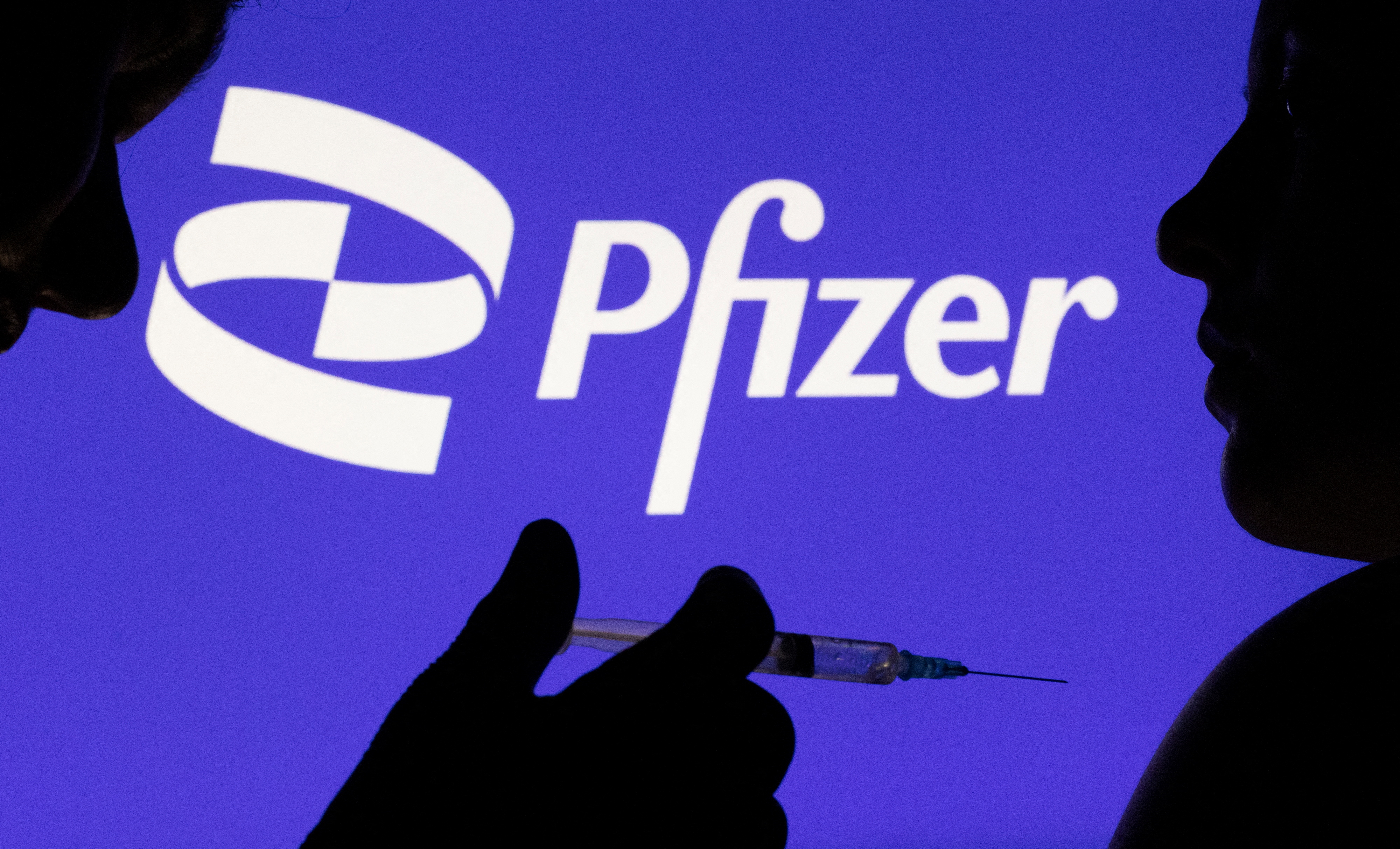 People pose with syringe with needle in front of displayed Pfizer logo