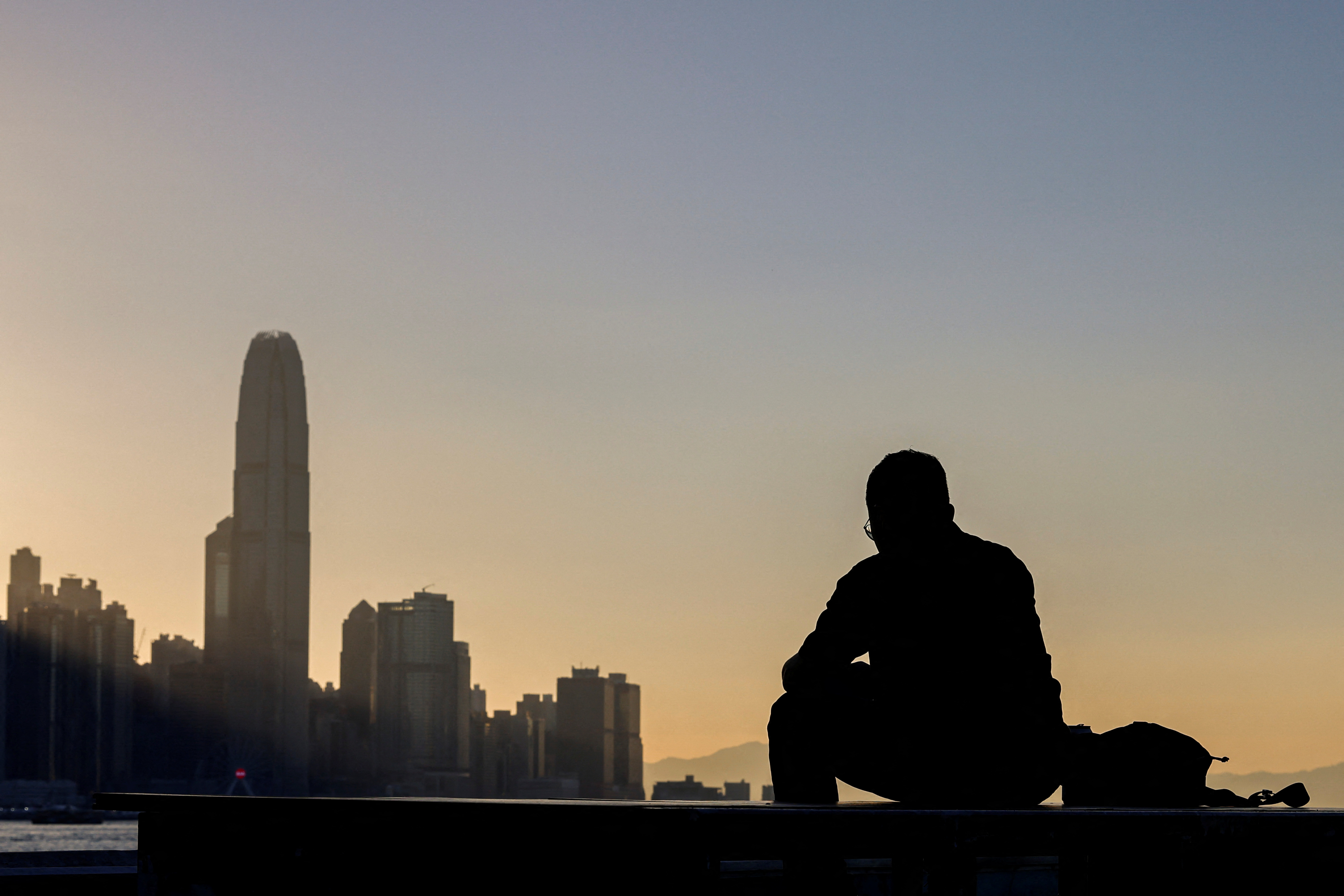 A man sits by the waterfront with the skyline buildings across Victoria Harbor in Hong Kong