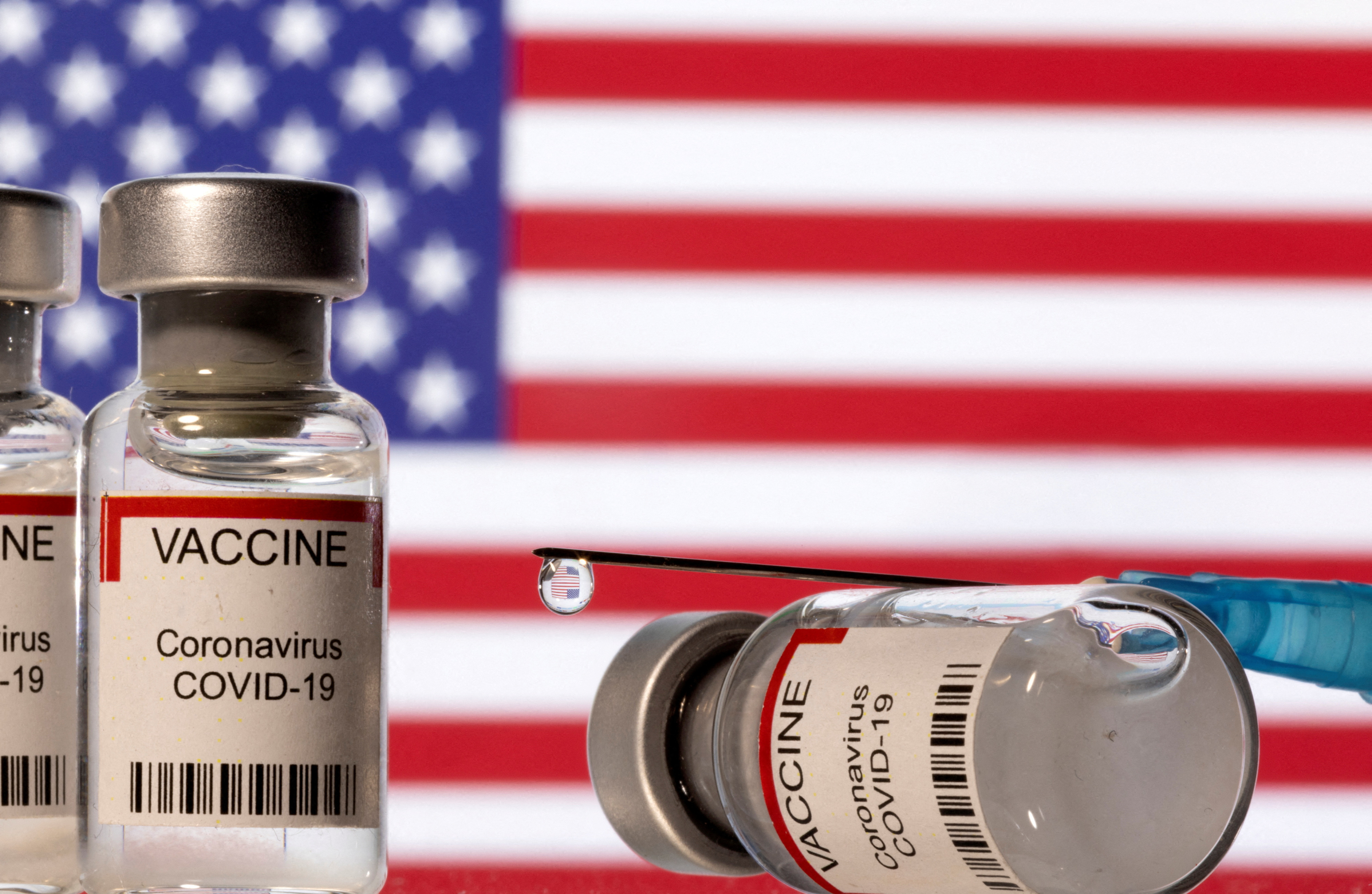 Illustration shows vials labelled "VACCINE Coronavirus COVID-19" and a syringe in front of a displayed U.S. flag
