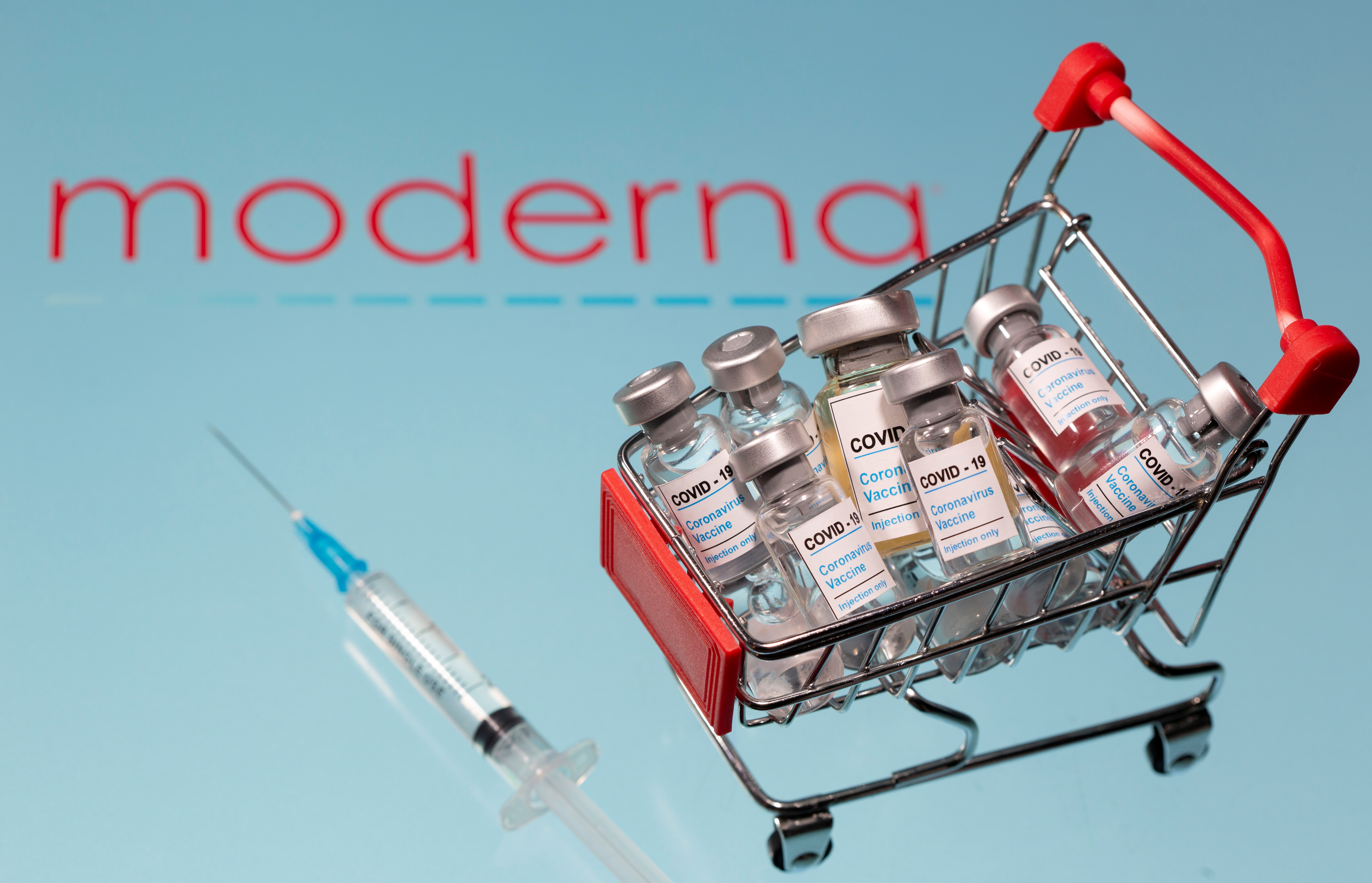 A small shopping basket filled with vials labelled "COVID-19 - Coronavirus Vaccine" and a medical syringe are placed on a Moderna logo