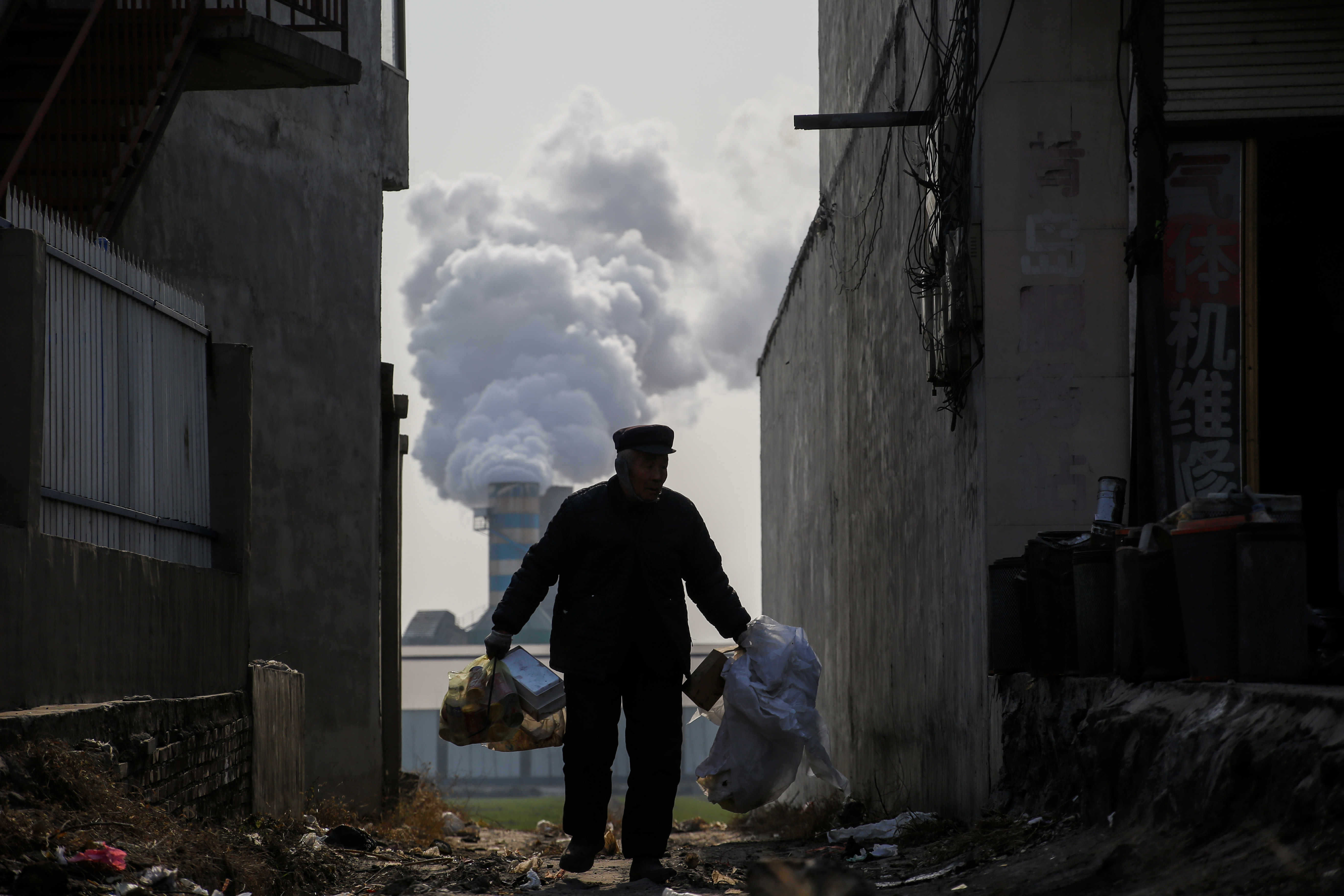 A man collects recyclables from an alley as smoke billows from the chimney of a factory in rural Gaoyi county near Shijiazhuang